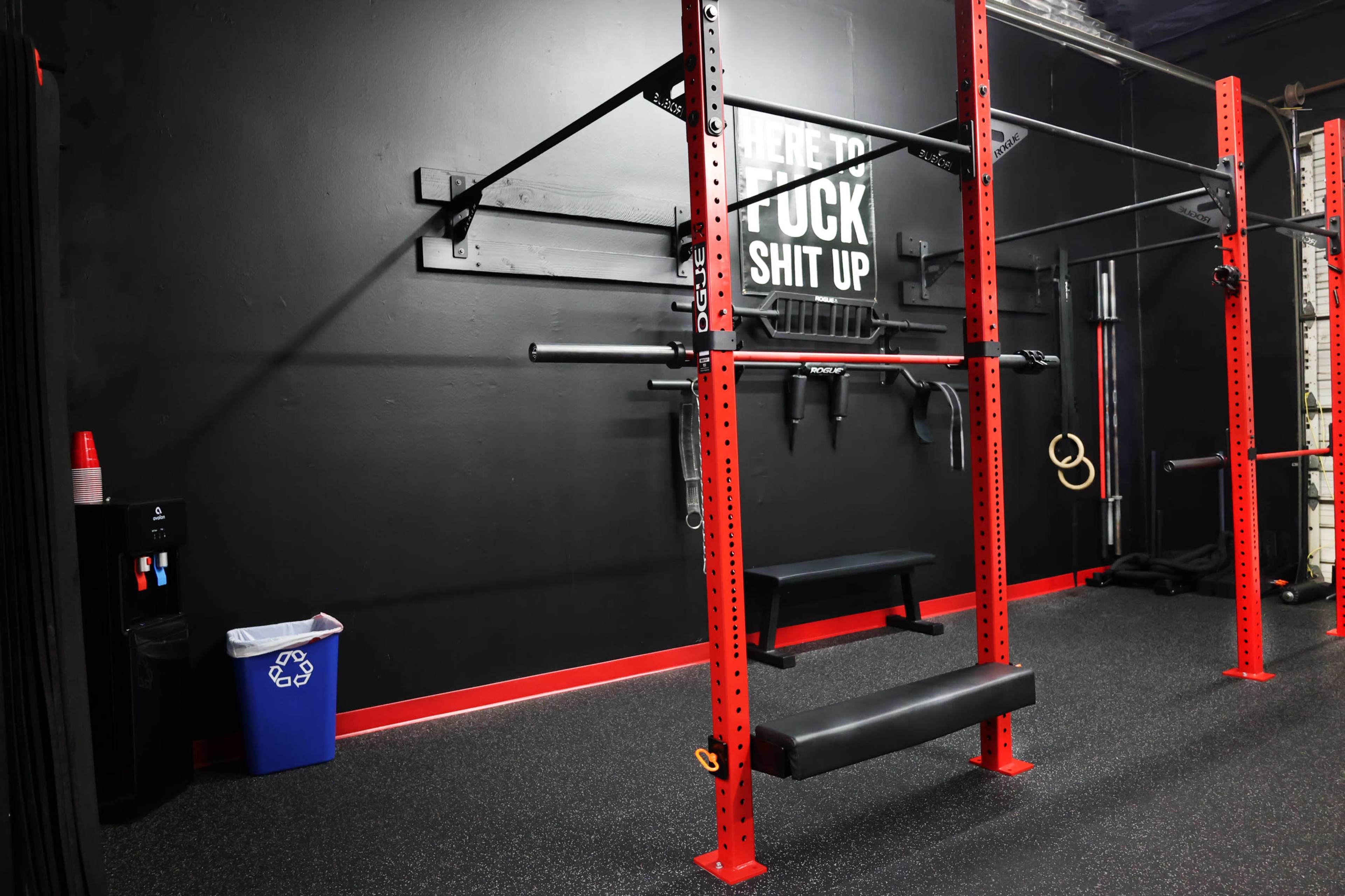 The image shows a section of a gym featuring red workout equipment, including a squat rack and benches, against a black wall with motivational text.