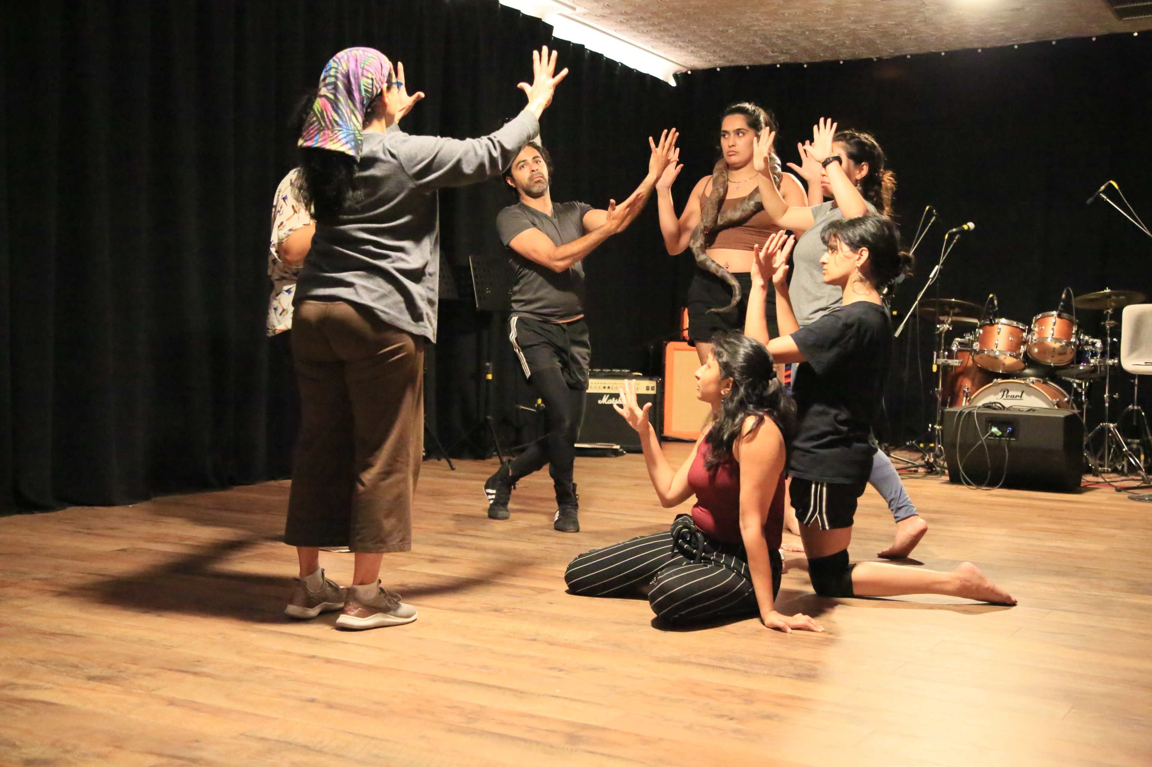 A group of six performers engages in an expressive movement exercise on a wooden stage with musical instruments in the background.