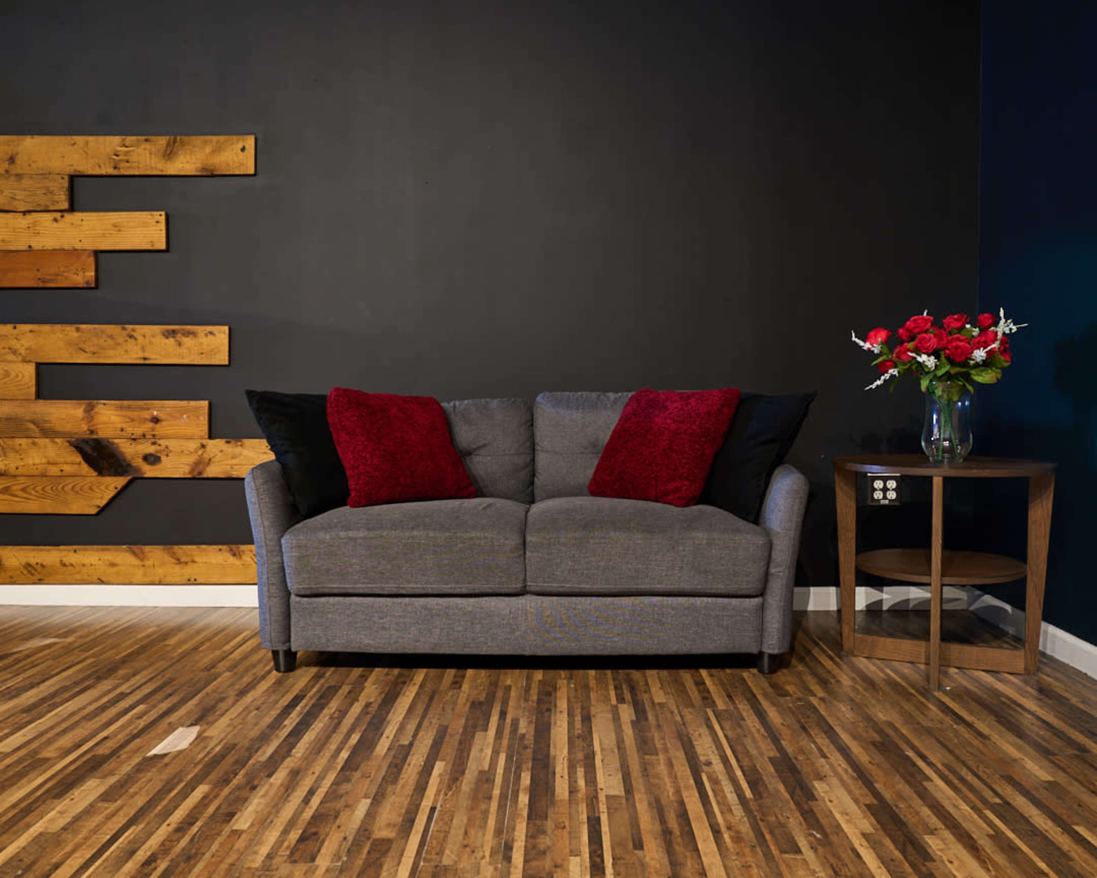 A gray couch with red and black pillows is positioned against a dark wall, complemented by a wooden wall art piece and a small table with a vase of flowers.