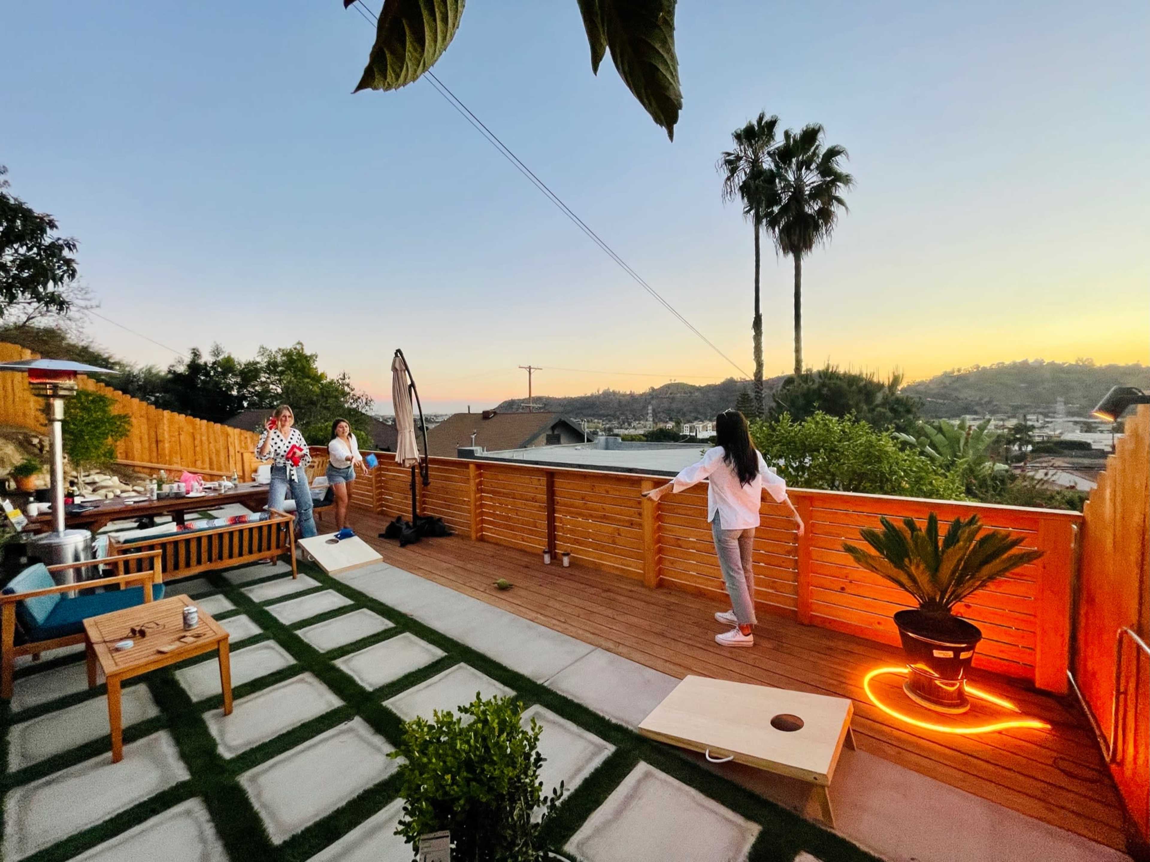 A backyard deck features people playing cornhole as the sun sets behind palm trees.