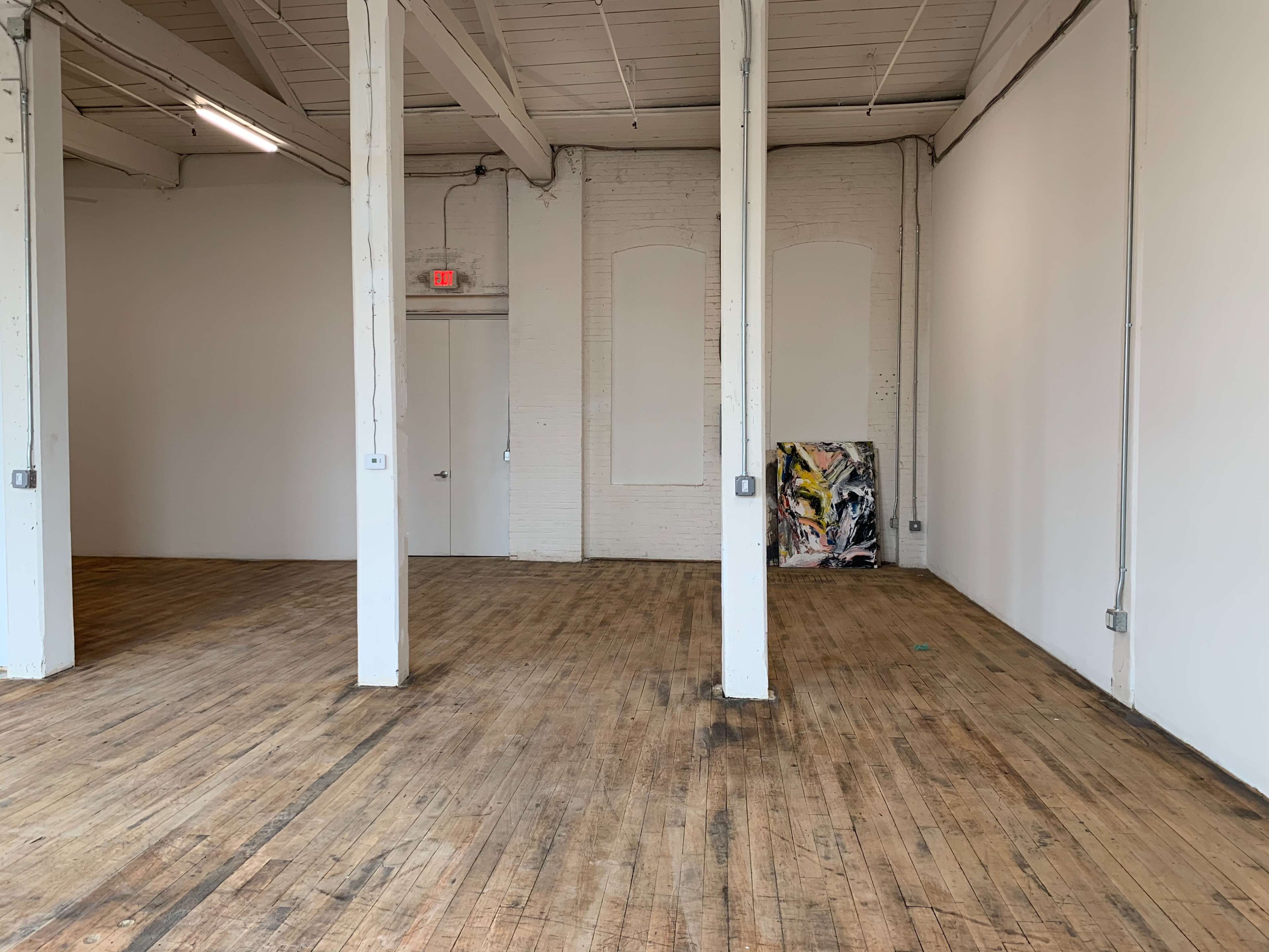 An empty art studio with wooden floors, white walls, and a few pillars supporting the ceiling, along with a painting in the corner.