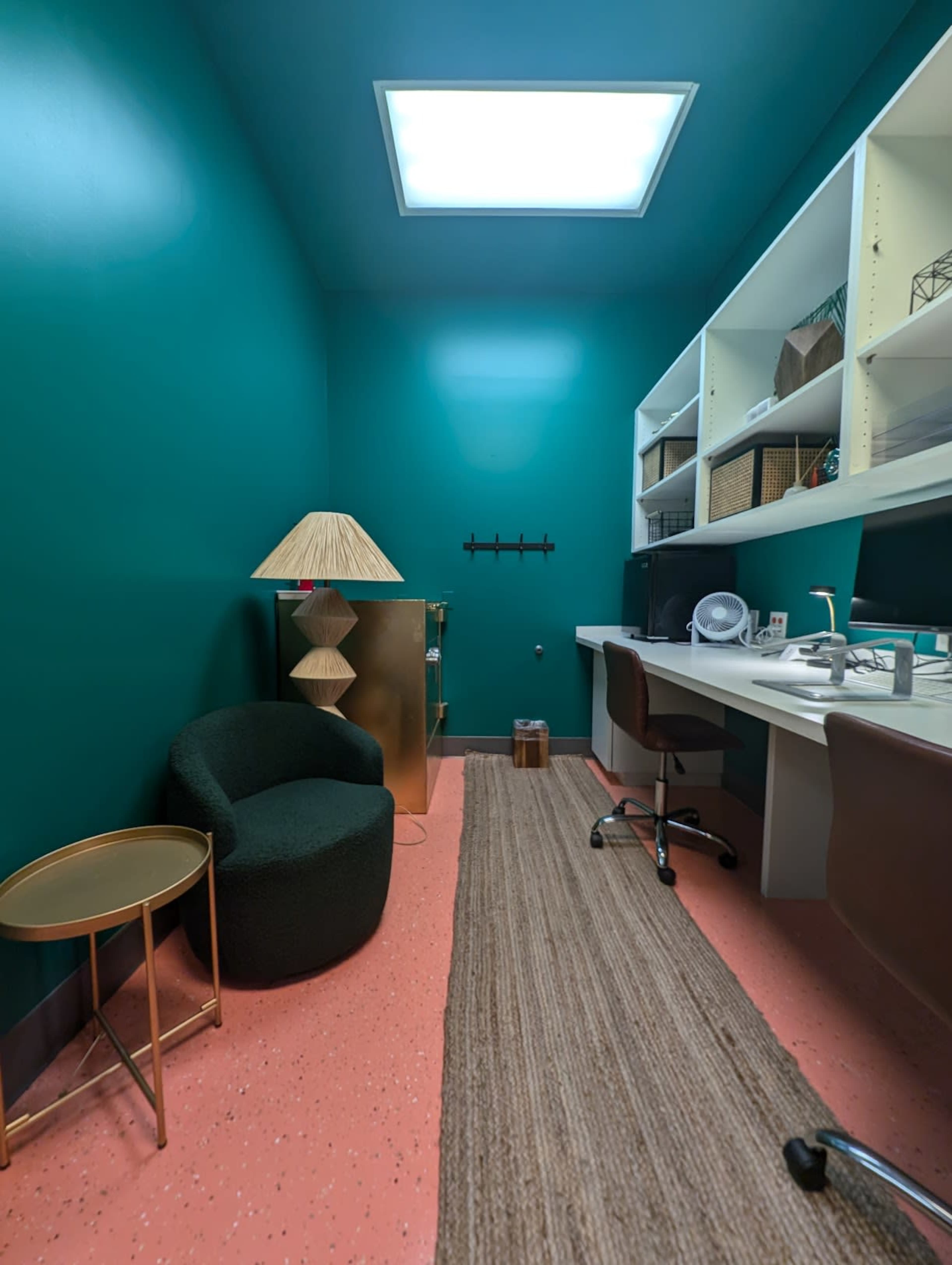 The image shows a small room with teal walls, a skylight, a green chair, a desk with a computer, and shelves containing baskets and office supplies.
