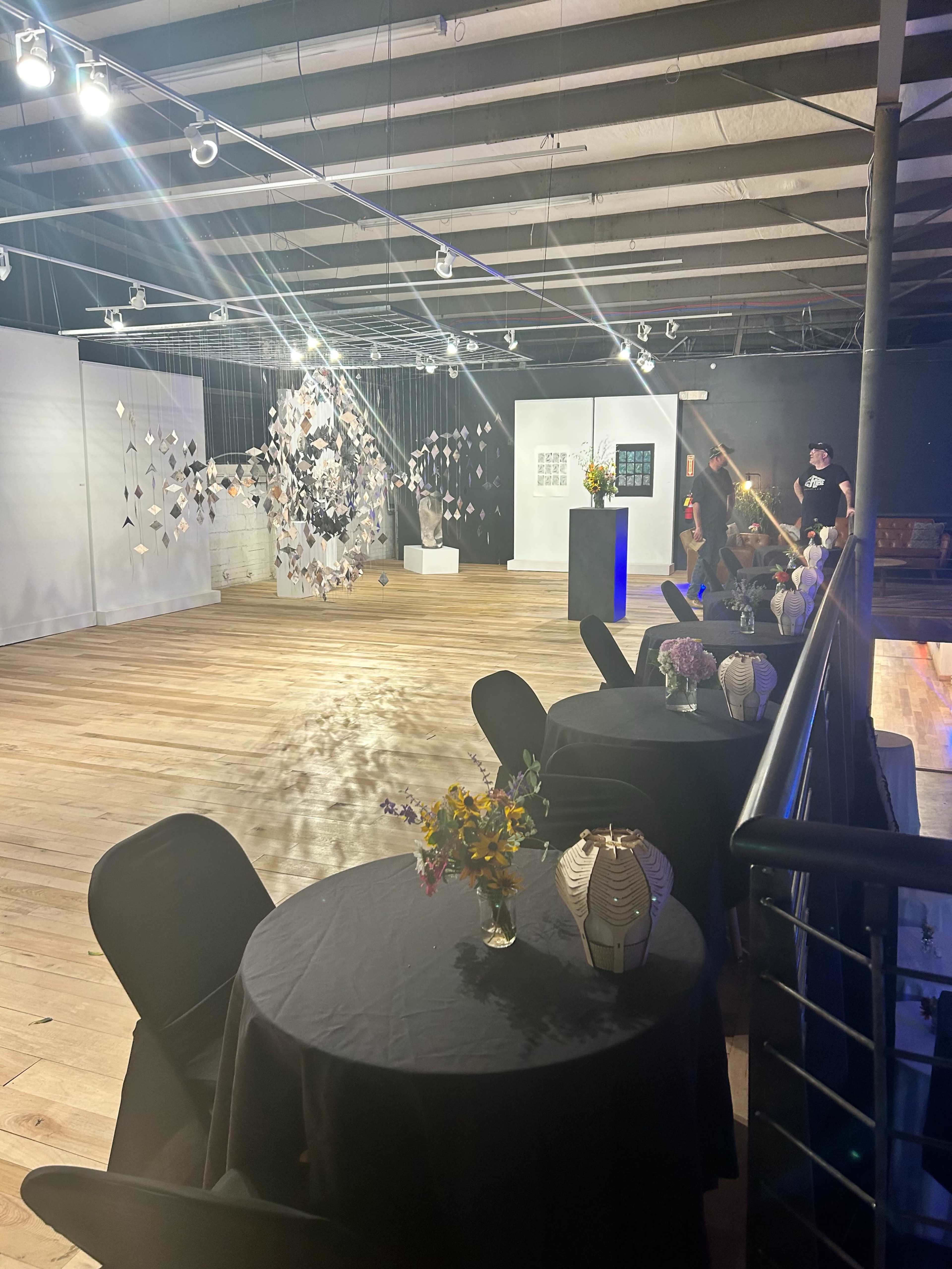 The image shows an art gallery featuring several tables with black tablecloths, sculptures, and hanging art installations, all reflected in a spacious wooden-floored area.