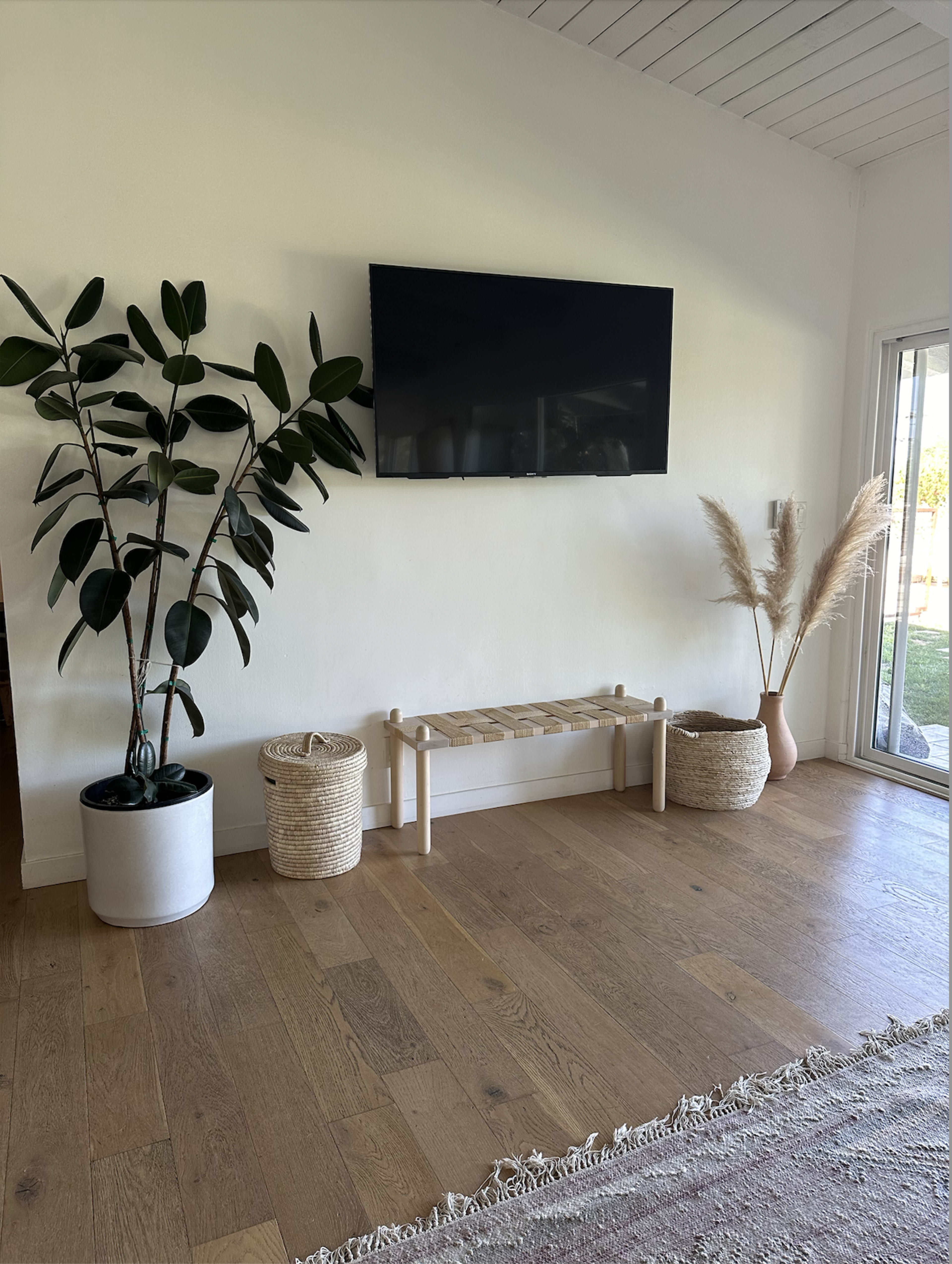 A minimalist living room with a television mounted on a white wall, alongside a tall potted plant, a woven bench, and decorative baskets.