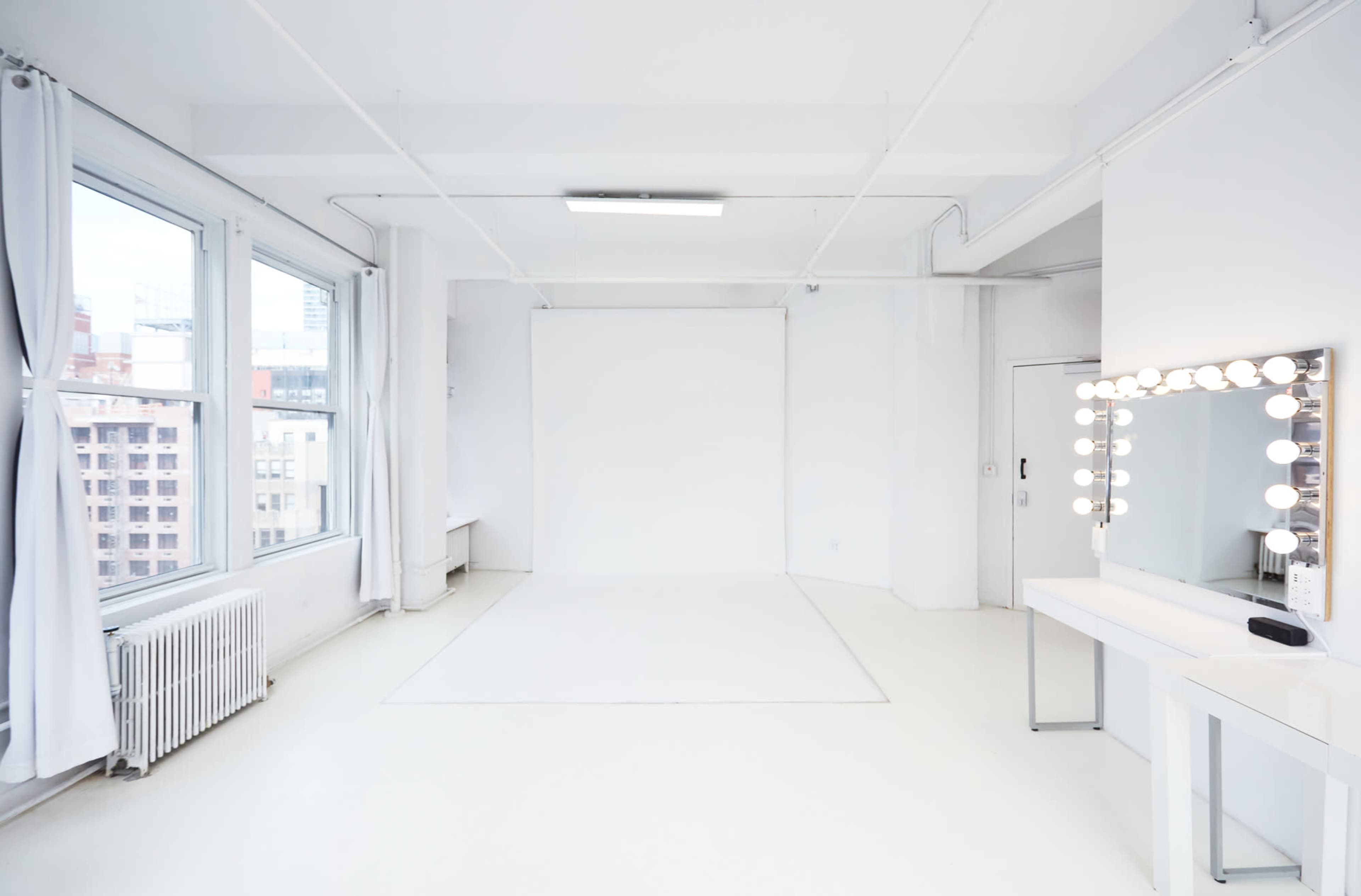 A bright, minimalist studio features white walls, a seamless backdrop, large windows, and a vanity mirror with lights.
