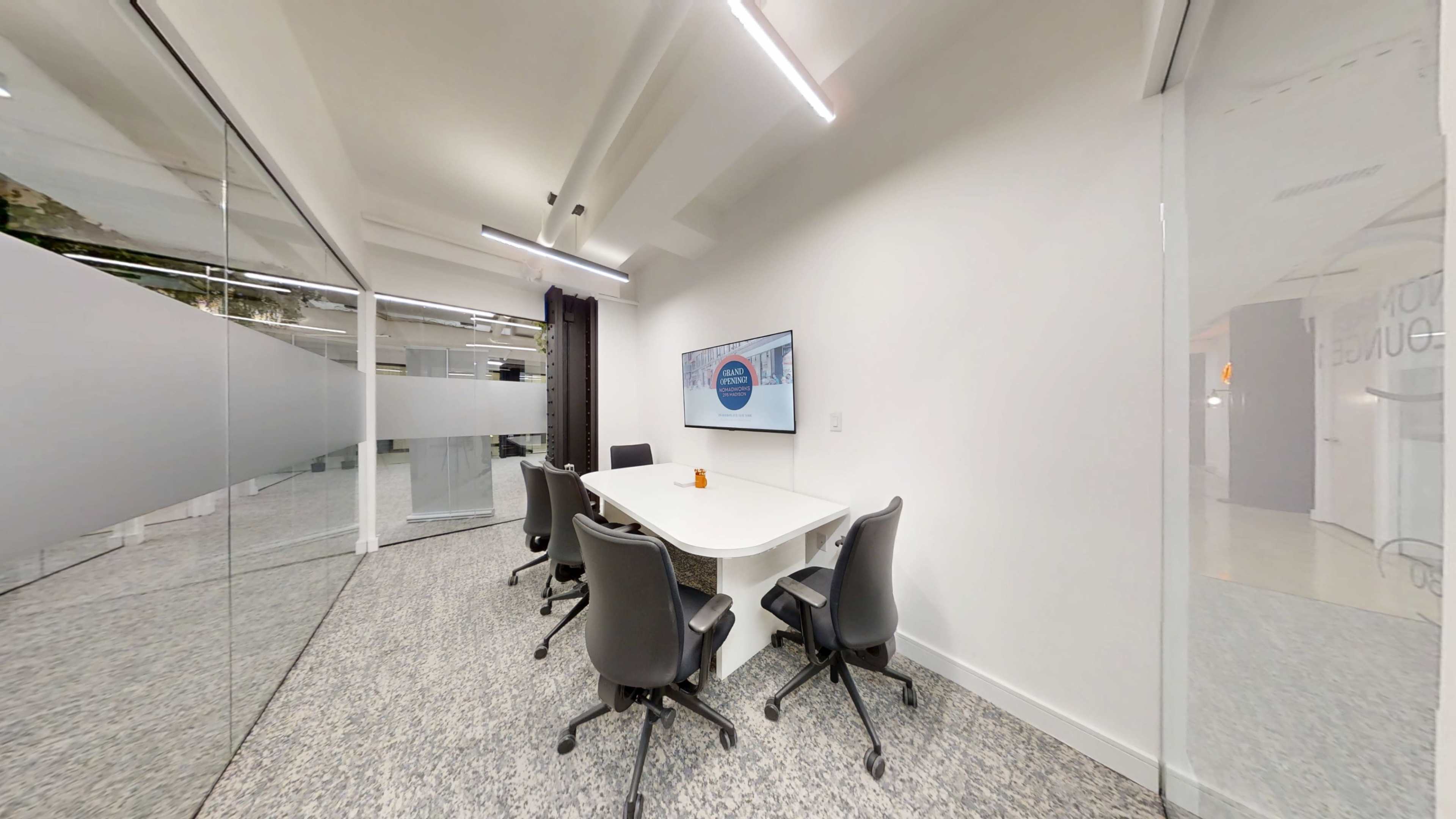 The image shows a modern conference room with a white table, four black chairs, and a wall-mounted screen displaying a presentation.