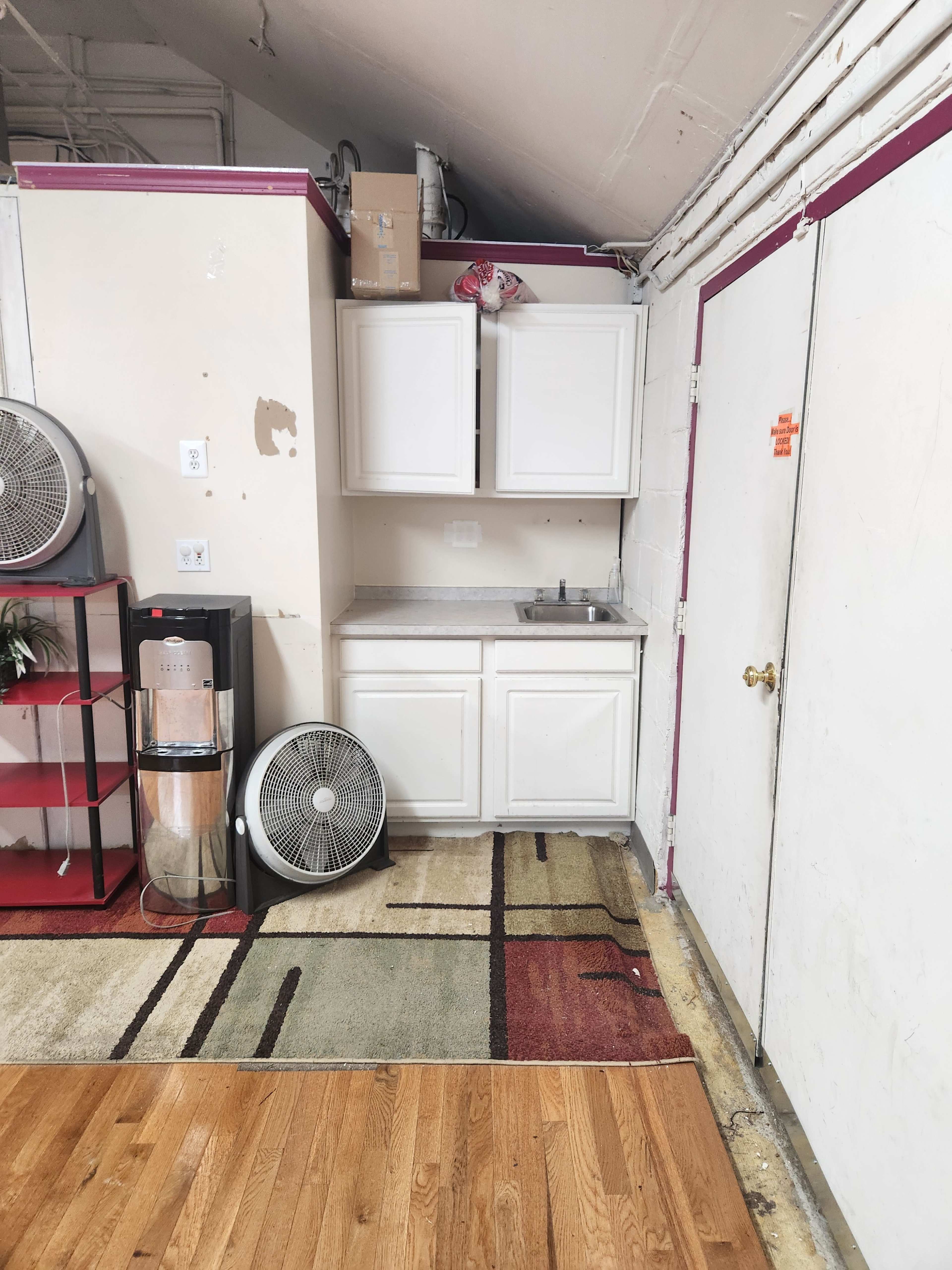 A small kitchenette corner with white cabinets, a sink, a standing fan, and a door, situated in a room with wooden flooring and a patterned rug.