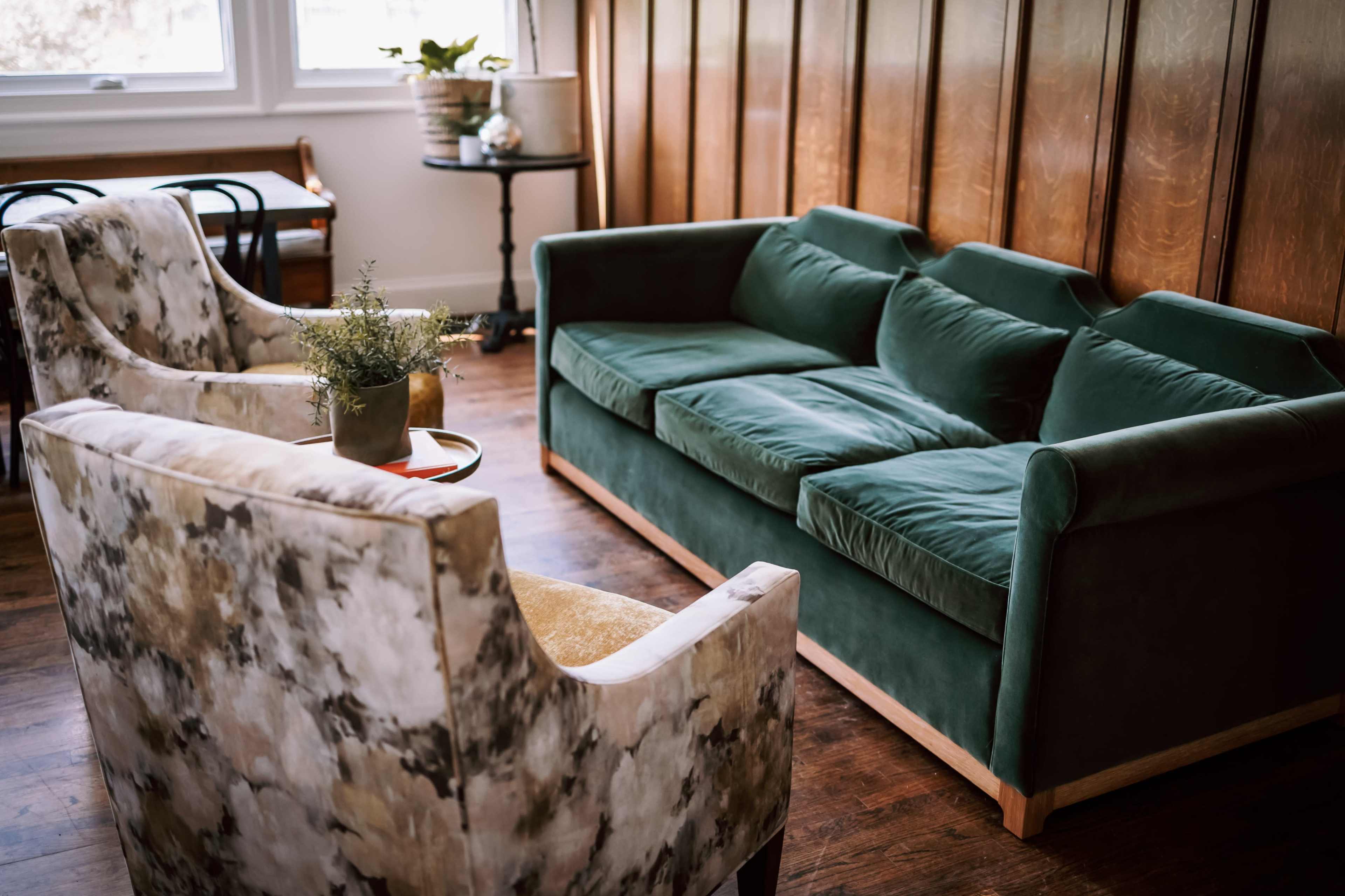 A green sofa and two patterned armchairs are arranged in a cozy living room with wooden wall paneling.