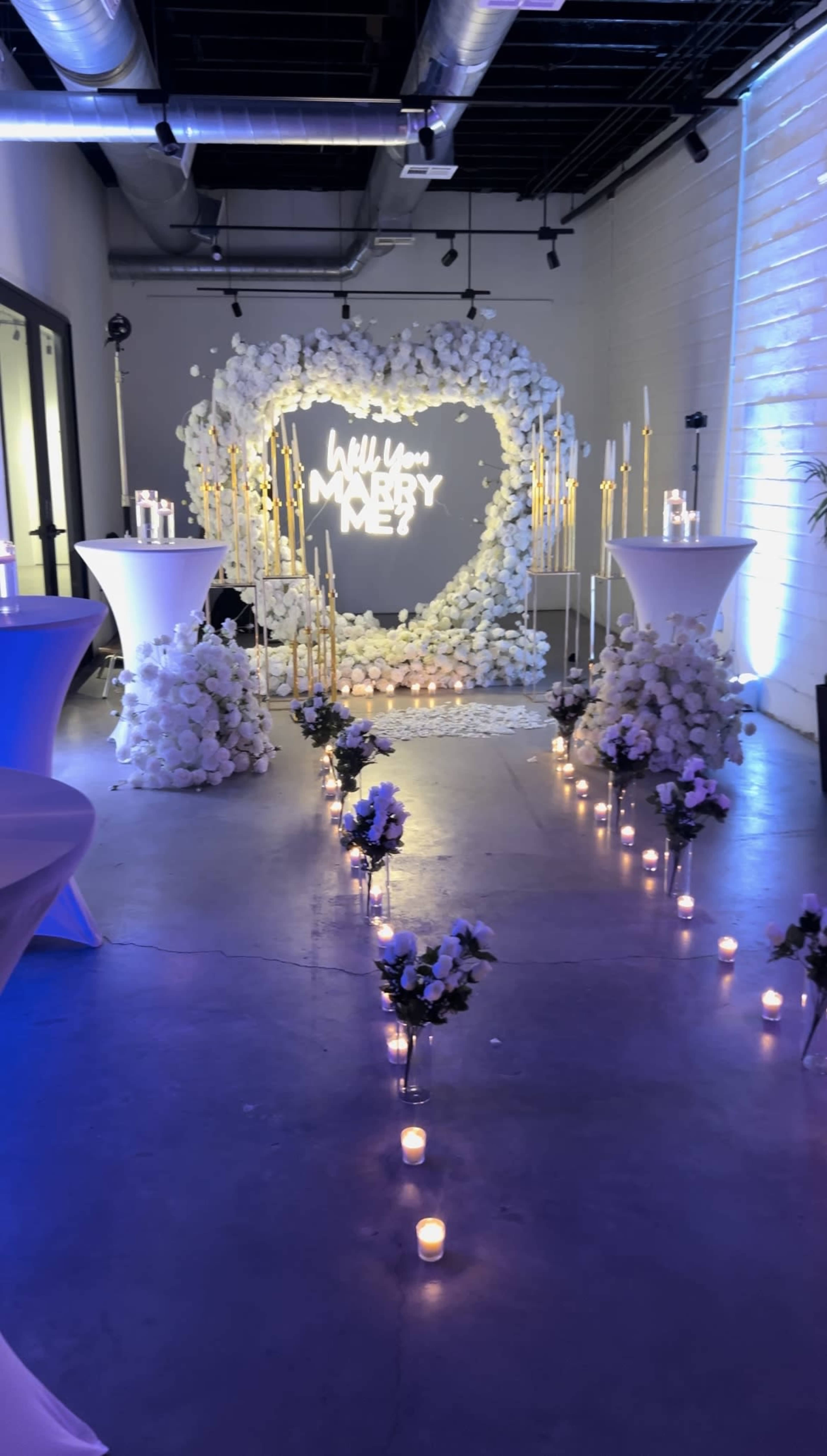 The image shows a decorated wedding proposal setup featuring a heart-shaped floral backdrop, illuminated signage, and candlelit pathways leading to the centerpiece.