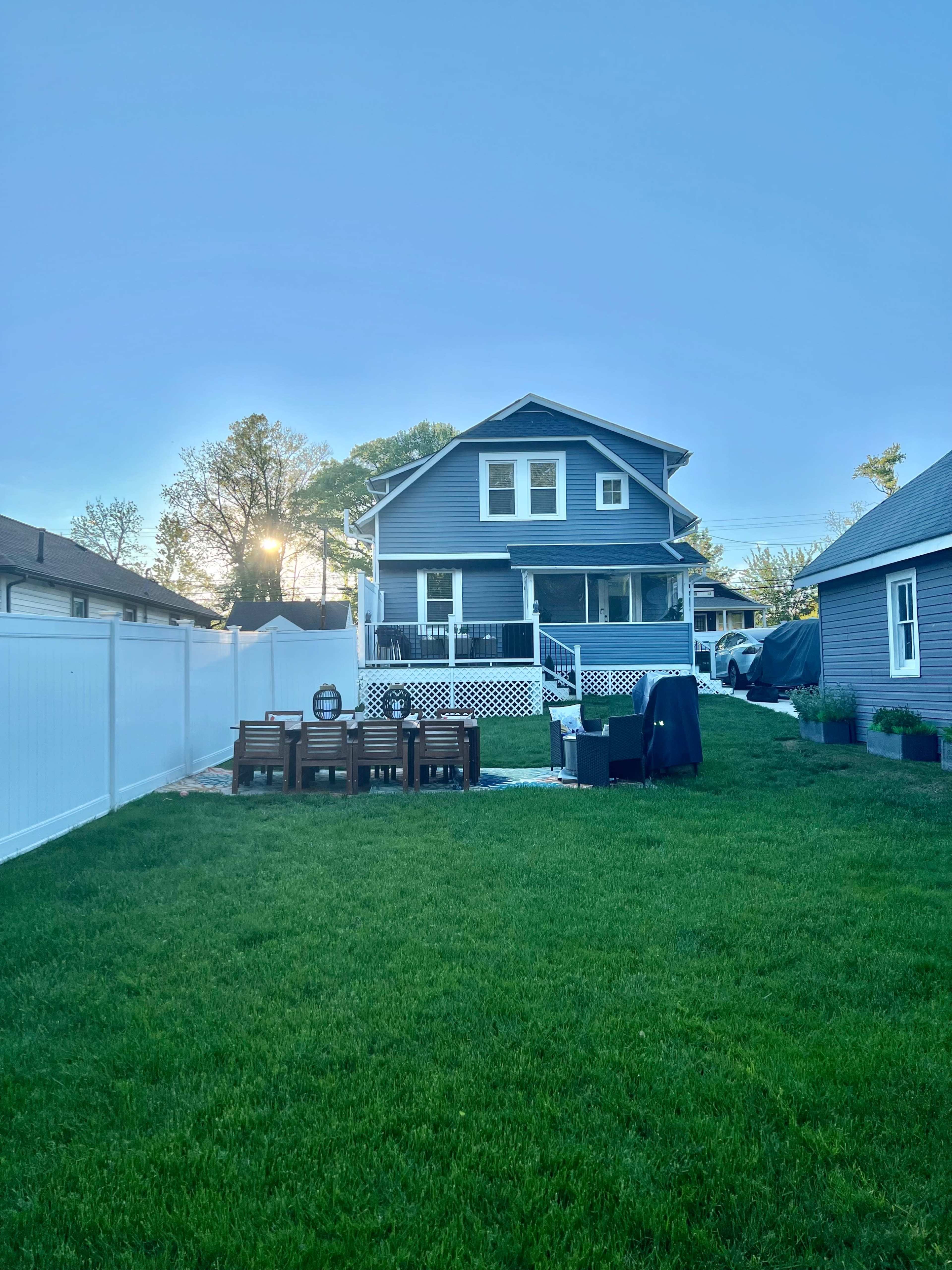 A blue two-story house with a porch overlooks a green lawn enclosed by a white fence, featuring a patio set in the foreground.