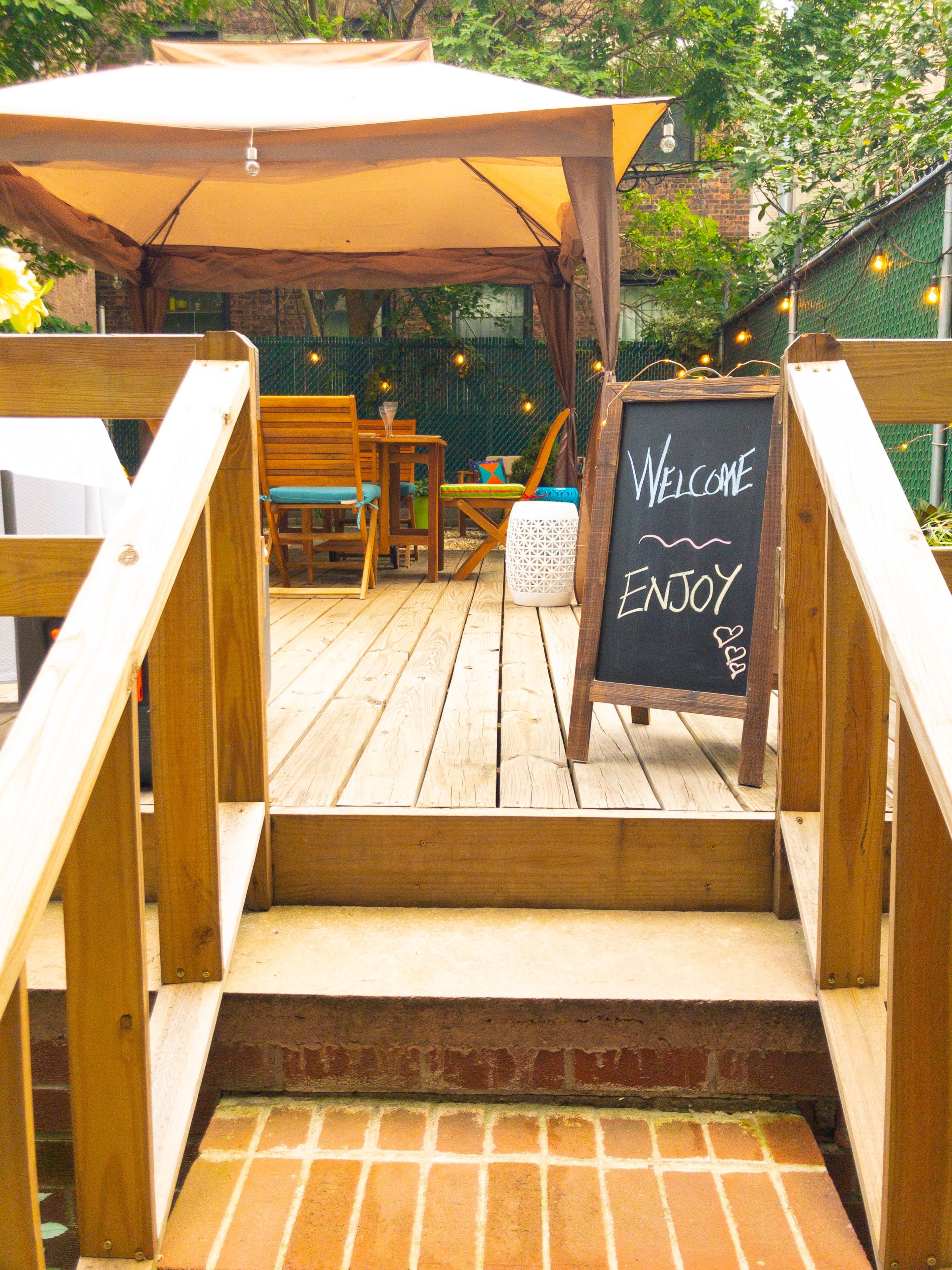 A welcome sign is positioned on a wooden deck that leads to a shaded seating area adorned with string lights.