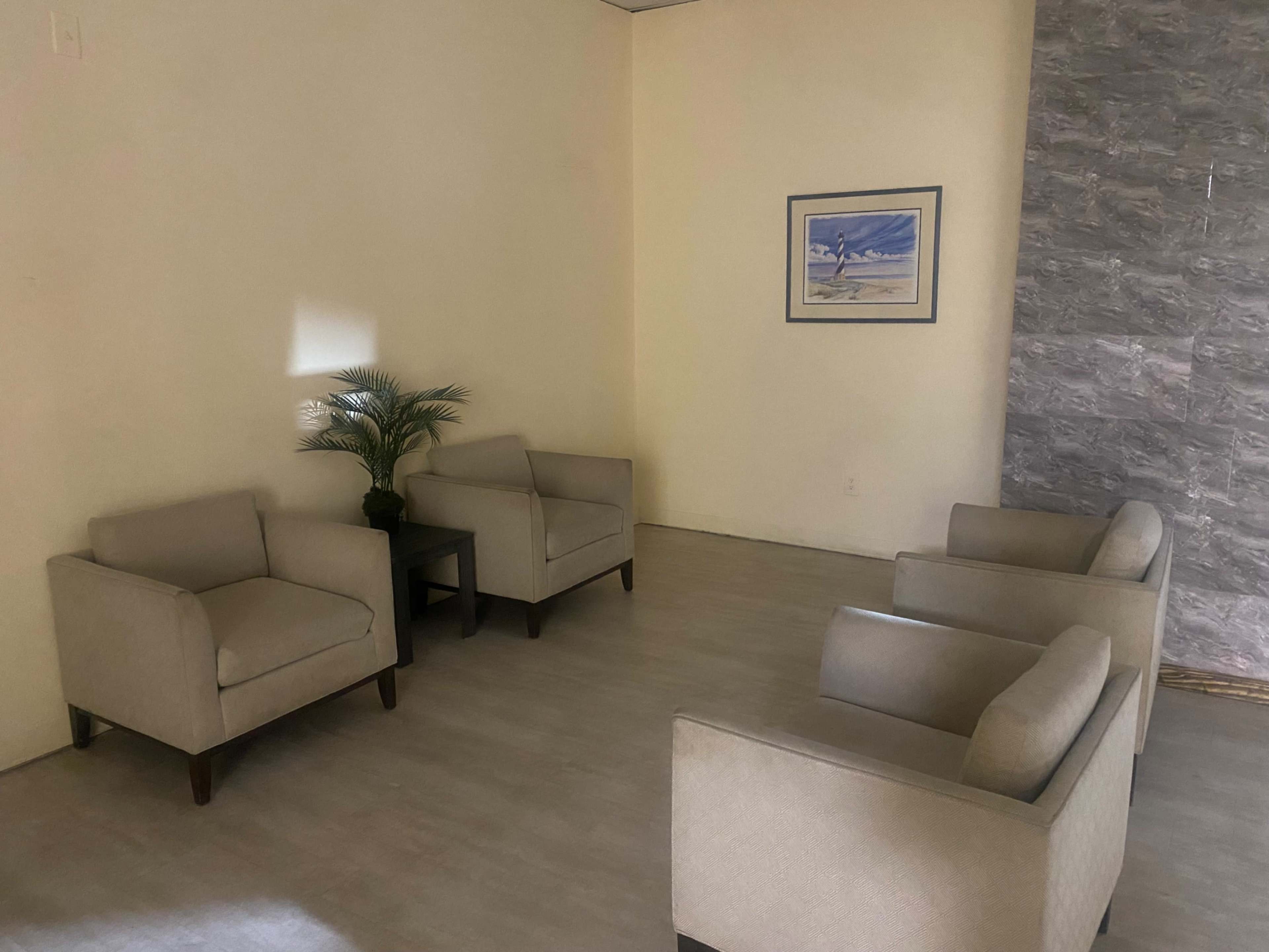 A waiting area features four armchairs arranged around a small table, with a potted plant against a light-colored wall and a framed picture on the opposite wall.