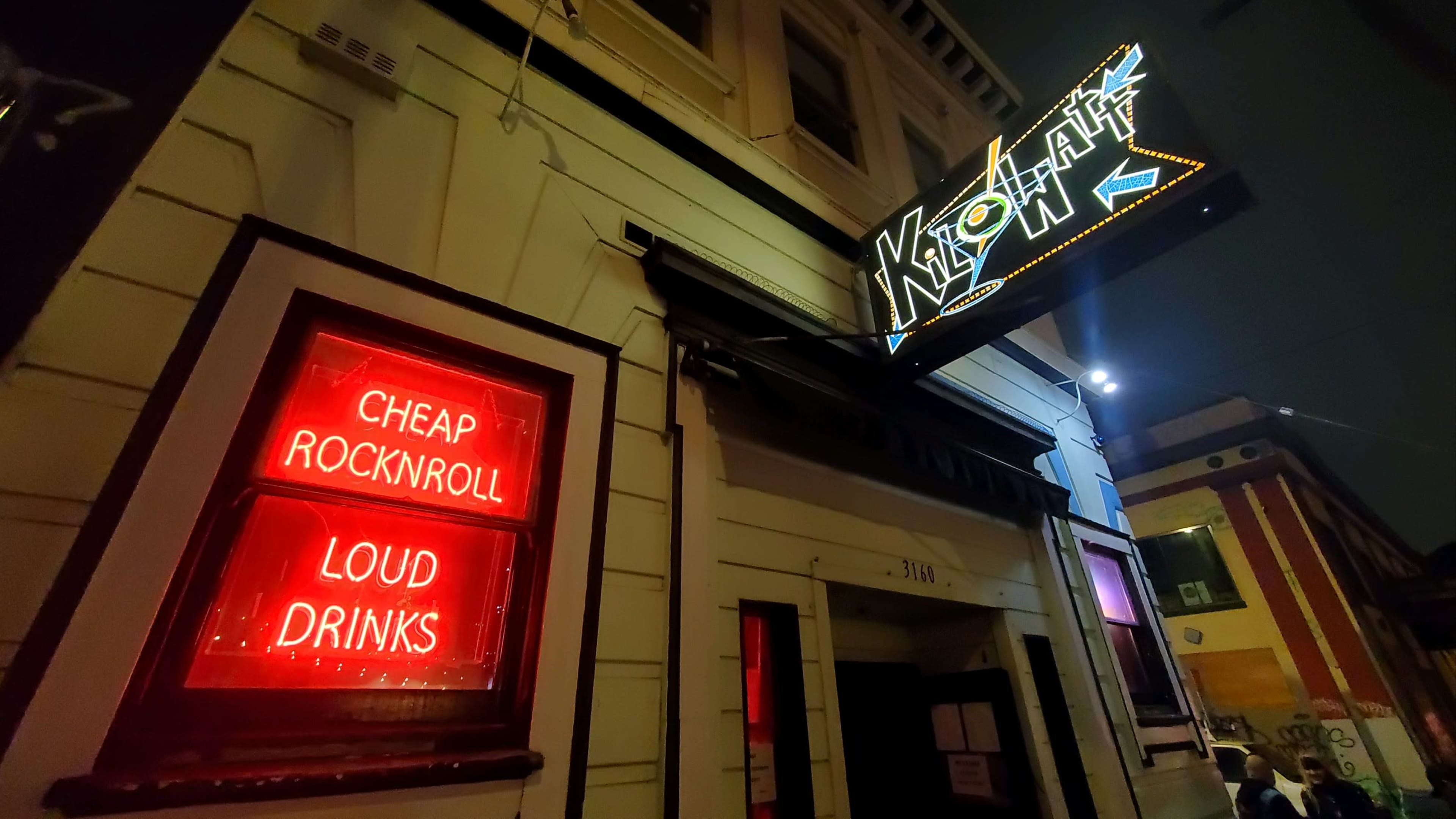 A bar entrance with a brightly lit sign and a window displaying a red neon advertisement for "Cheap Rocknroll" and "Loud Drinks."