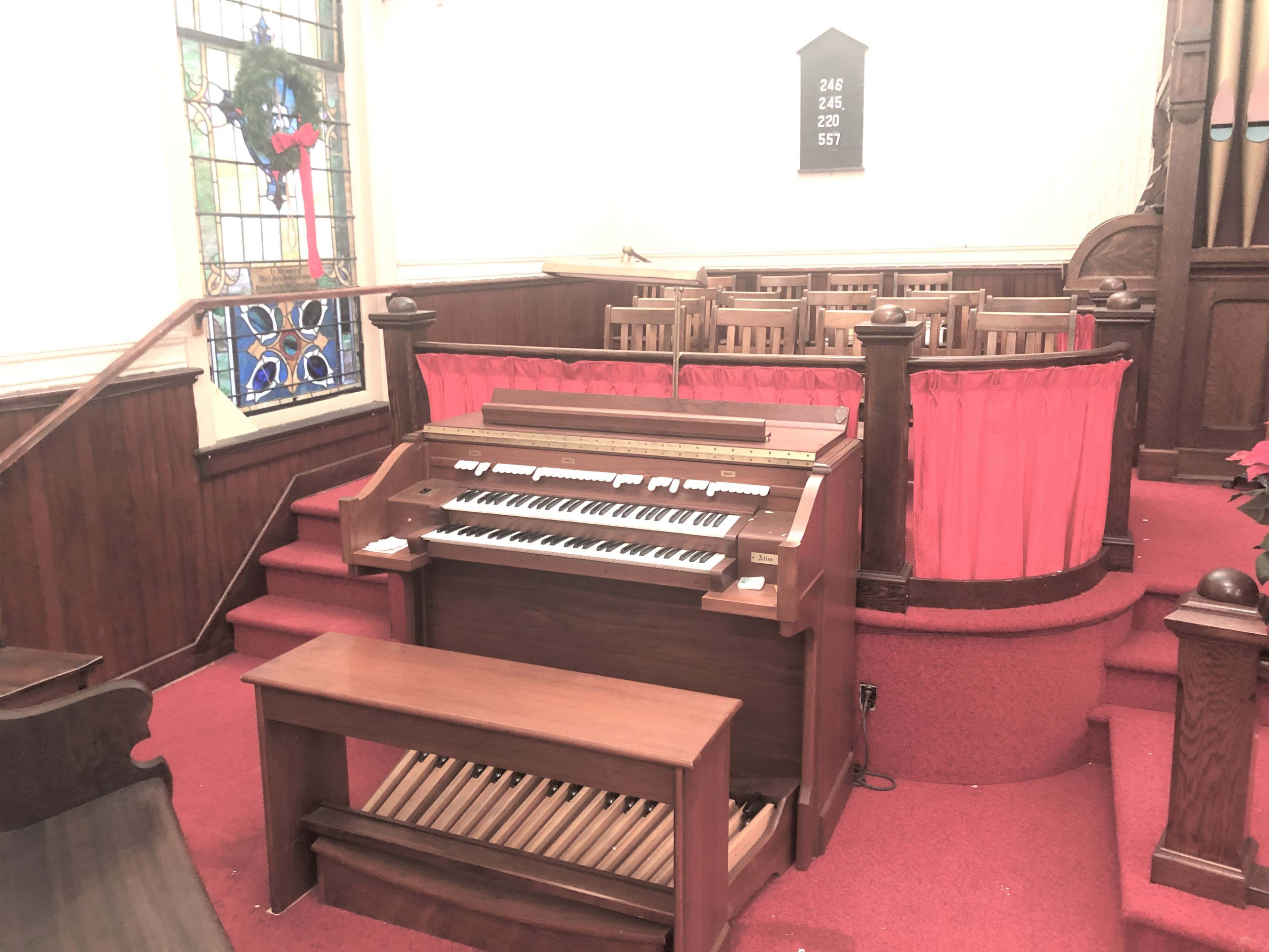 A wooden organ situated on a raised platform in a church, with red carpeting and a row of wooden pews in the background.