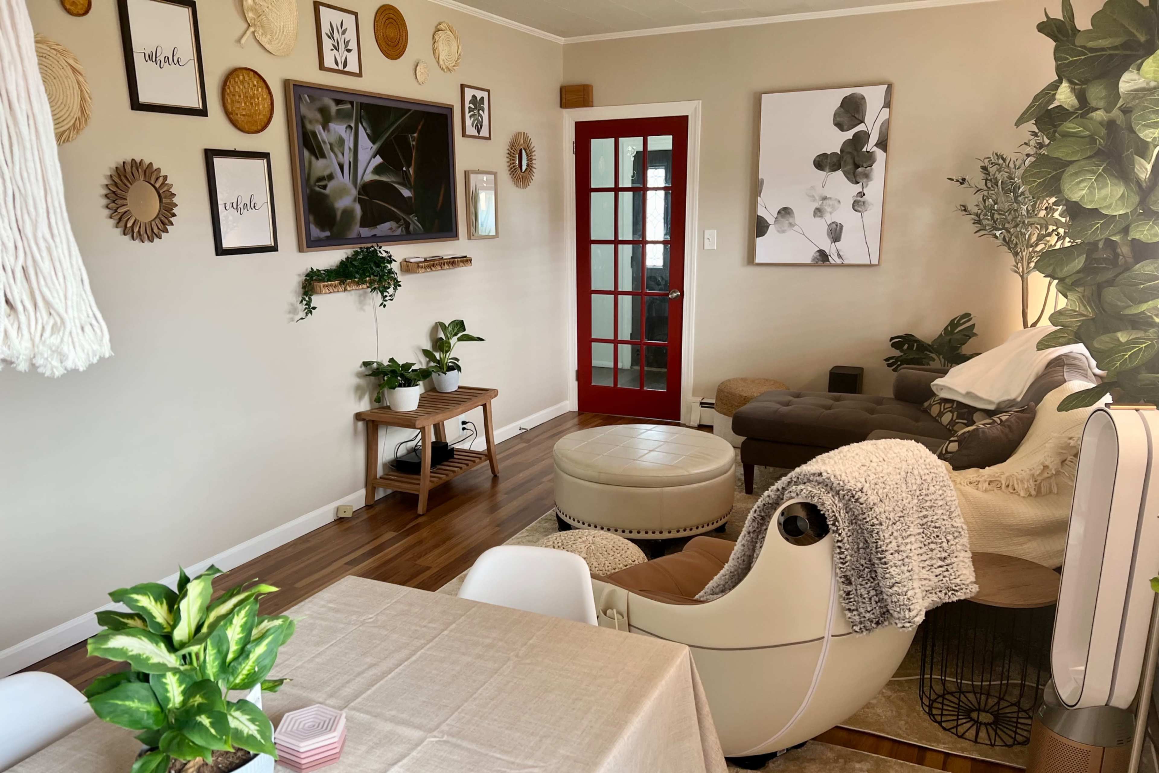A cozy living room features a mix of wall art, plants, a chaise lounge, and a red door leading to another space.