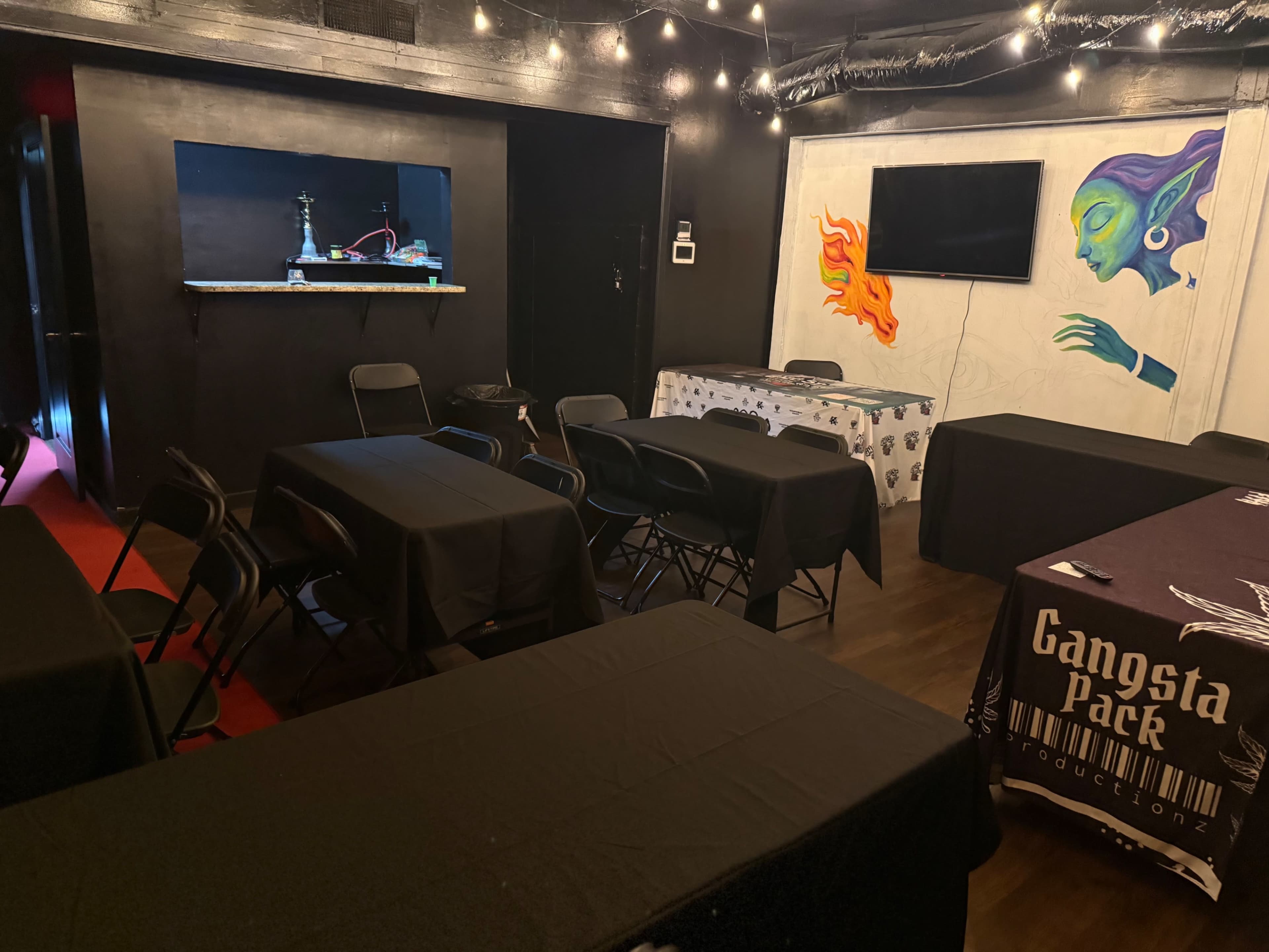 The image shows a dimly lit room with several round tables covered in black tablecloths, a TV on the wall, and a colorful mural of a face.