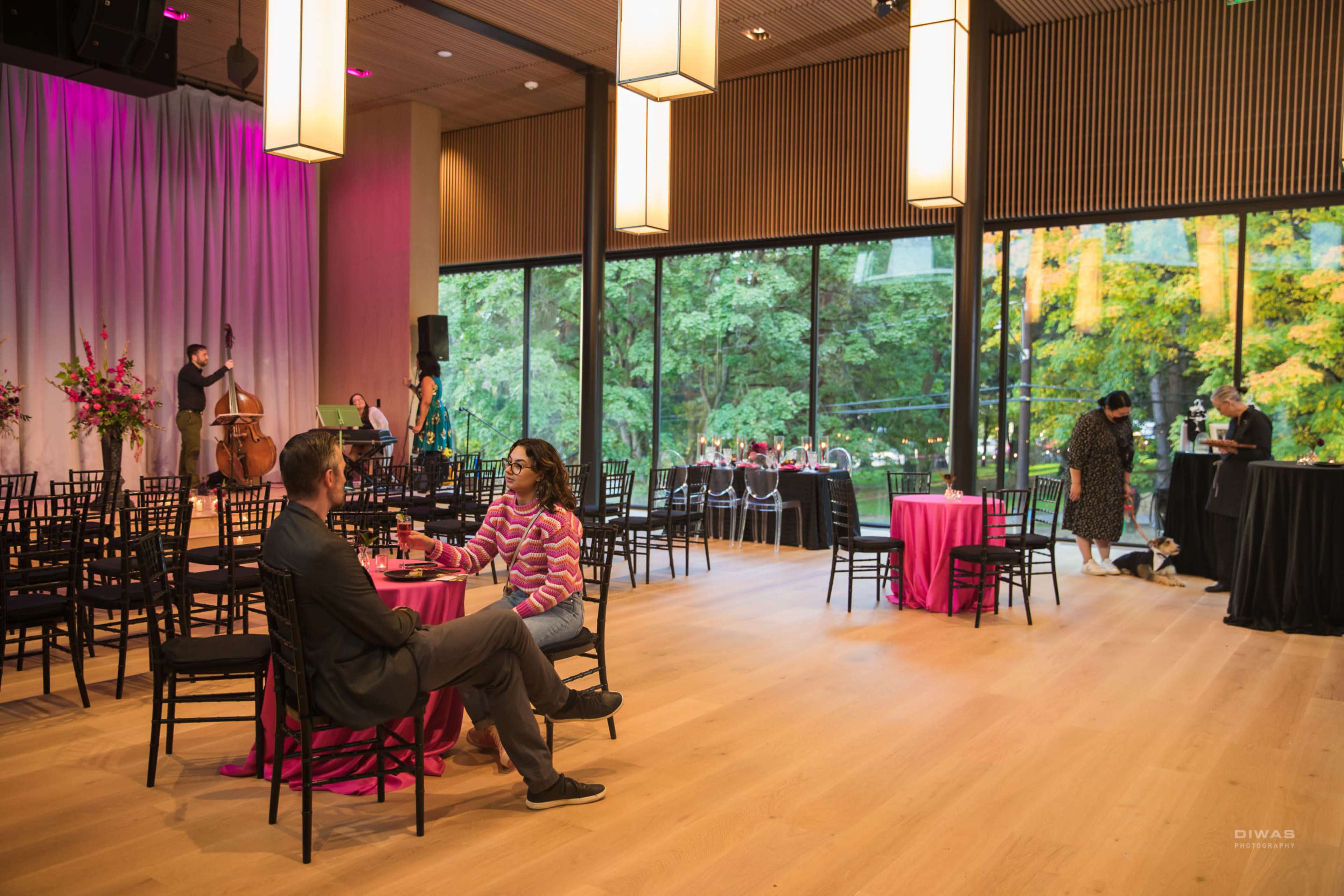 A couple sits at a table in a spacious venue with large windows overlooking a garden, while a small band performs in the background.