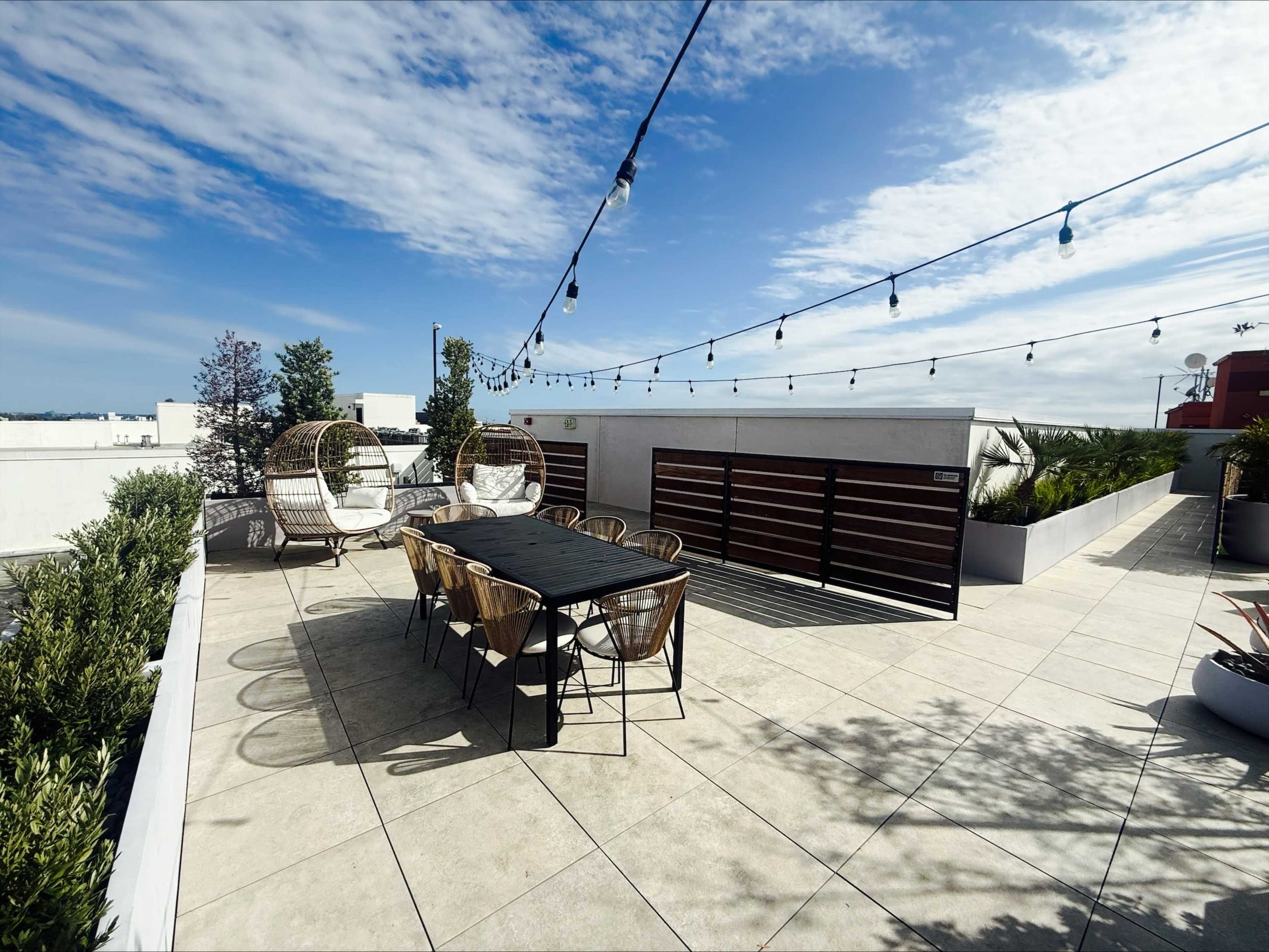 The image shows a rooftop terrace featuring a long dining table, six outdoor chairs, two hanging loungers, and decorative string lights above the area.