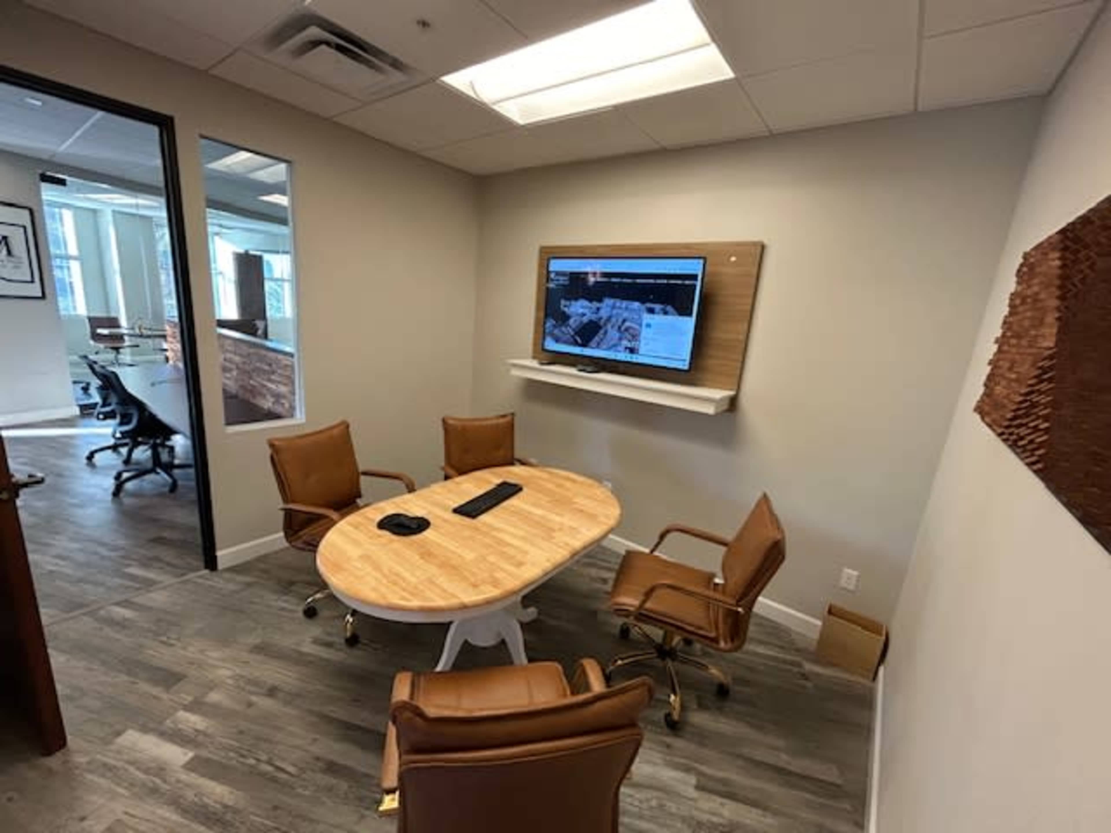 A small conference room features a circular wooden table surrounded by four chairs and a mounted television on the wall.