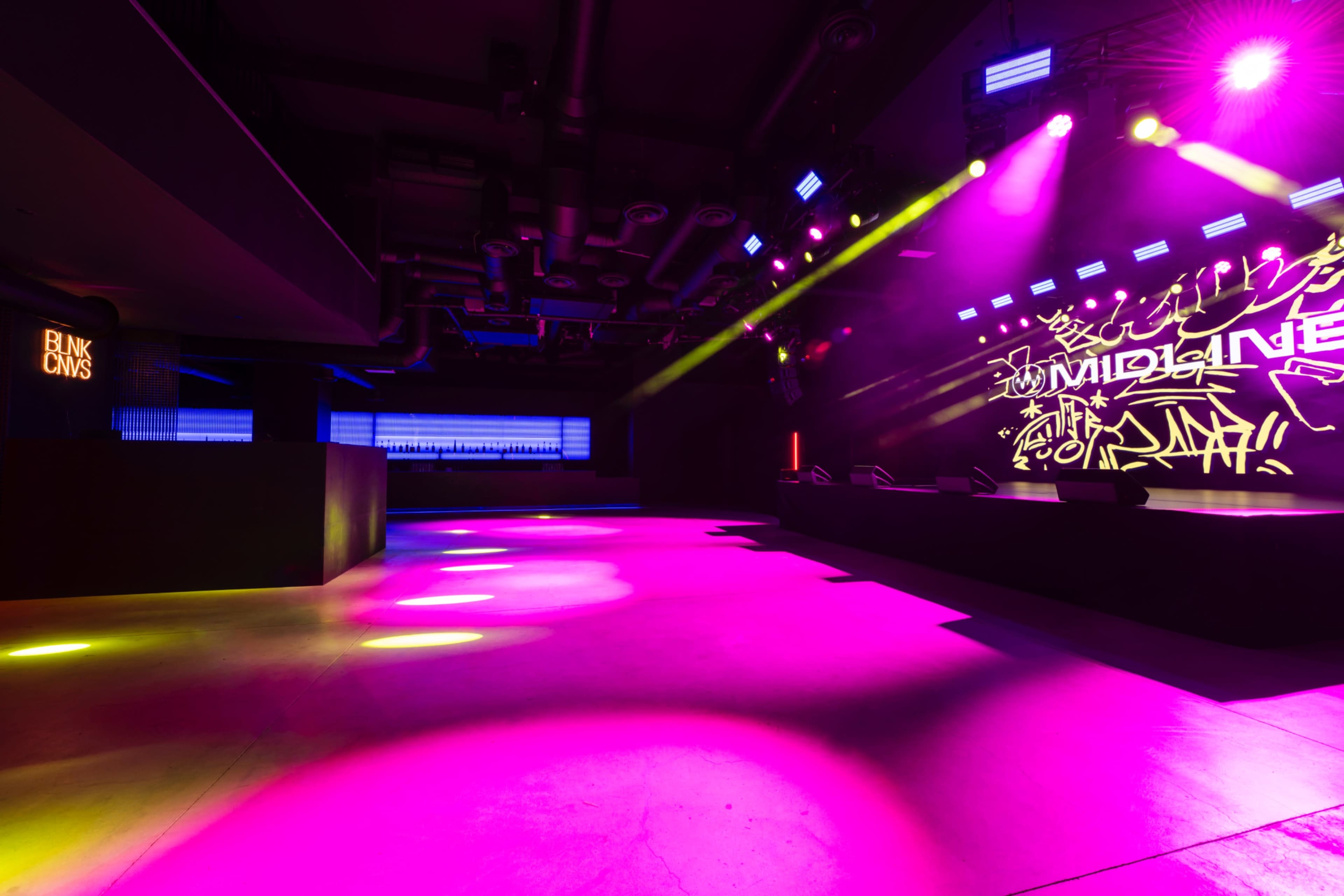 A brightly lit nightclub scene features a spacious dancefloor illuminated by colorful lights, with a DJ booth and graffiti-style visuals on the wall.