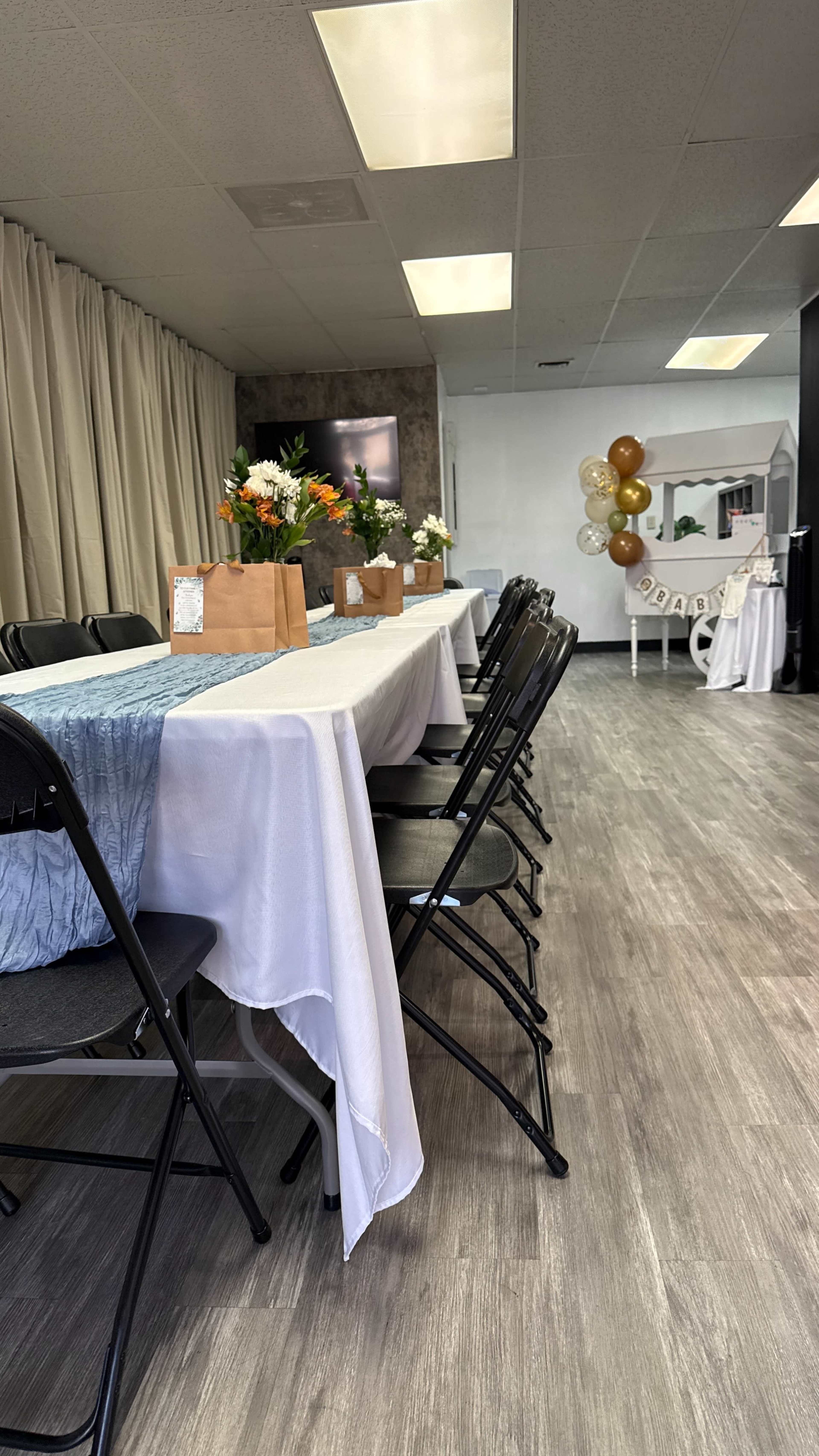 A long table is set with white and blue tablecloths, surrounded by black chairs, and decorated with flower arrangements and gift bags.