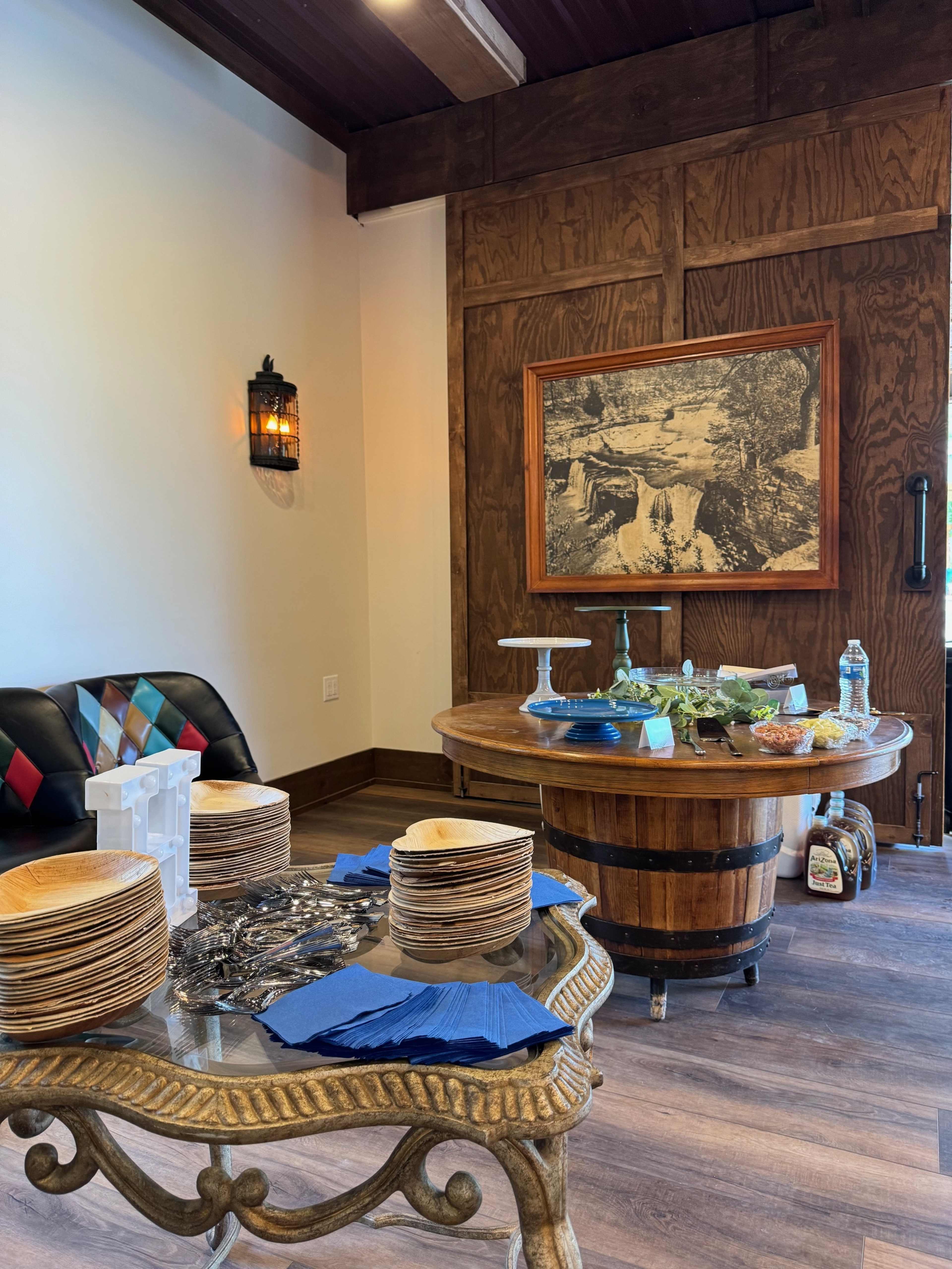 The scene features a wooden table with food arrangements, surrounded by stacked plates and a glass table displaying napkins and utensils, set in a well-lit room with rustic decor.