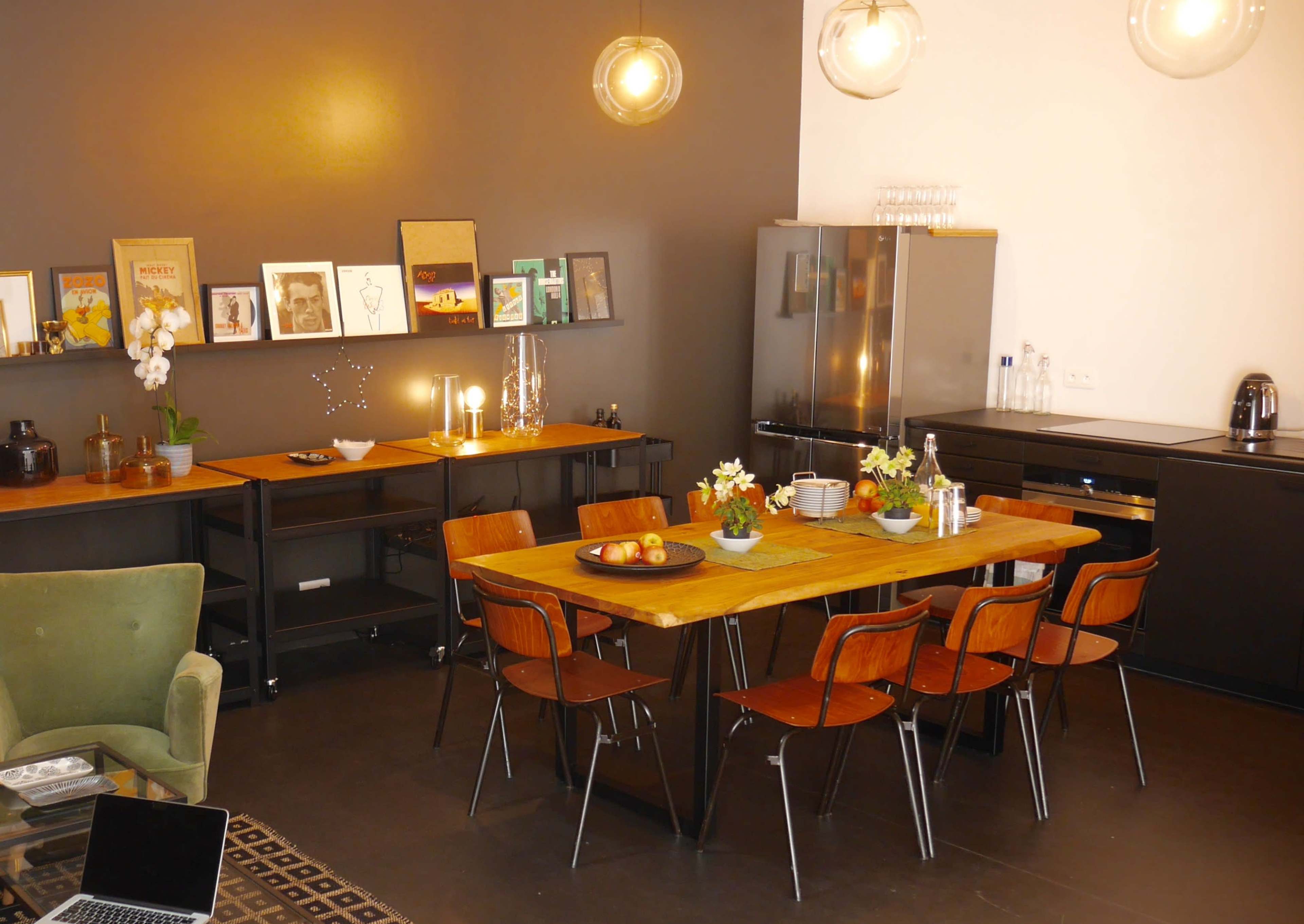 The image depicts a modern kitchen and dining area featuring a wooden table surrounded by chairs, with artwork displayed on the walls and a sleek refrigerator in the corner.