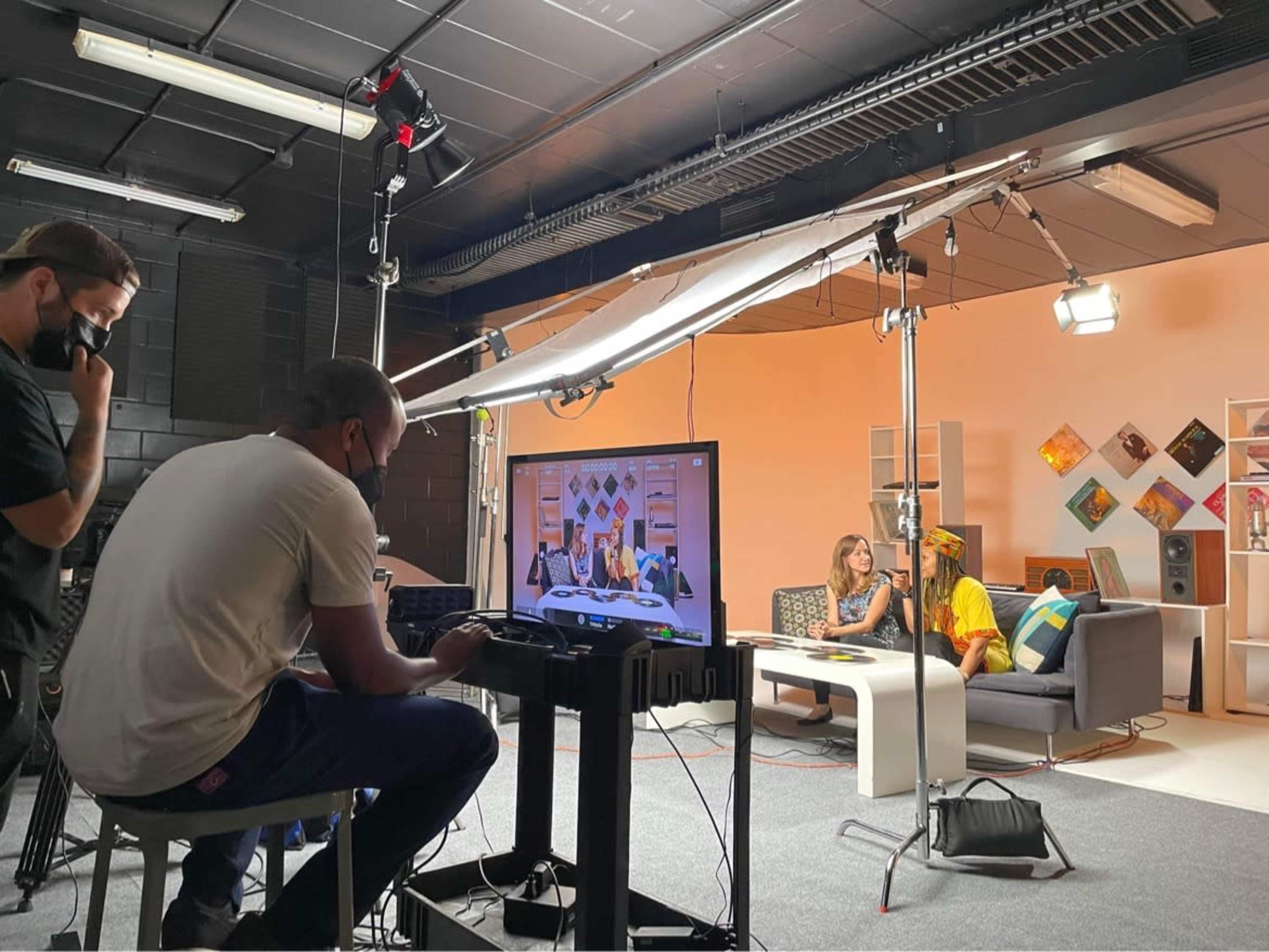 A television studio setup features two people engaged in conversation on a couch, while a camera operator and equipment are visible in the foreground.