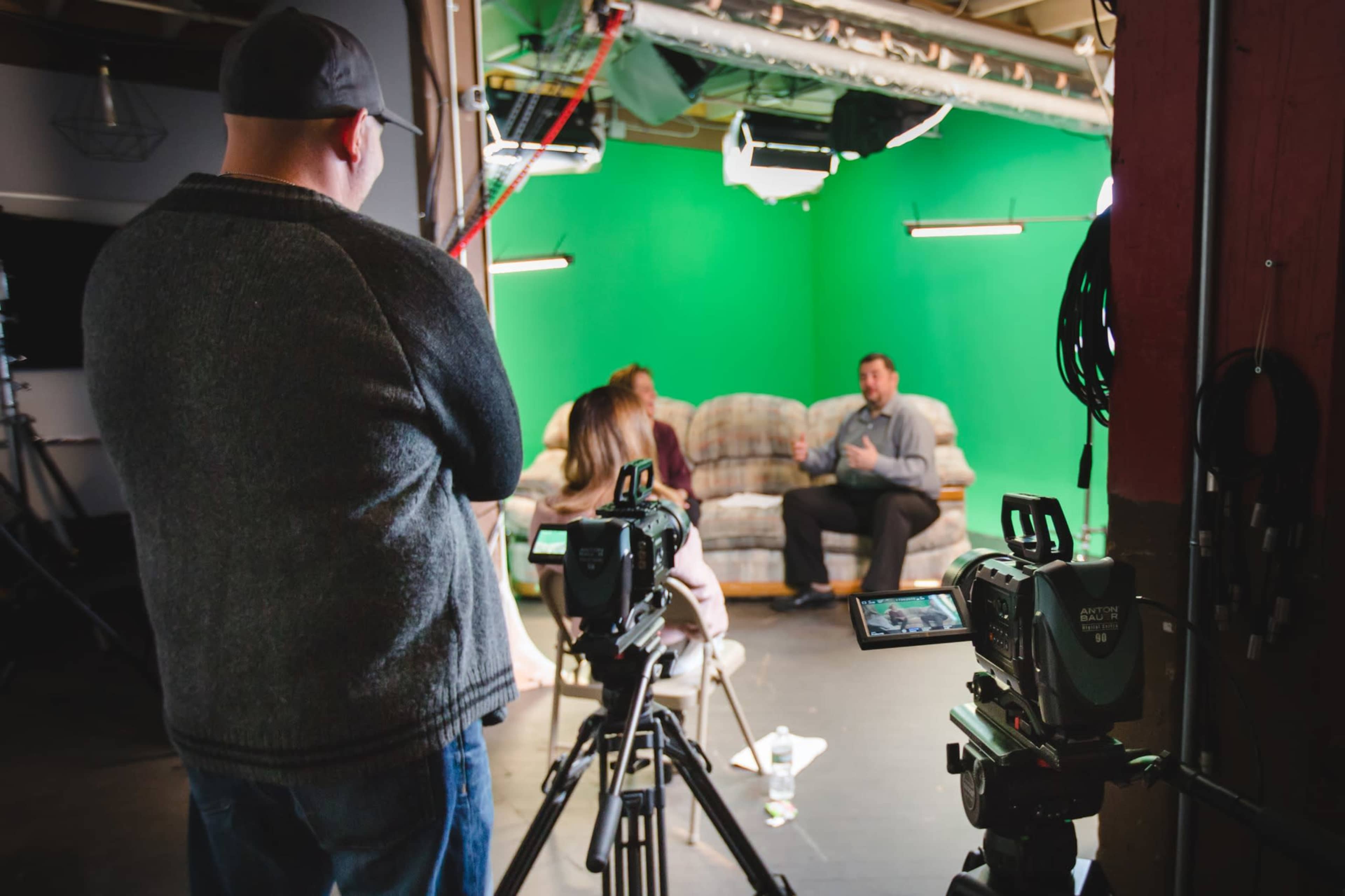 A man in a sweater operates a camera while a woman and a man are seated on a couch in front of a green screen during a video shoot.