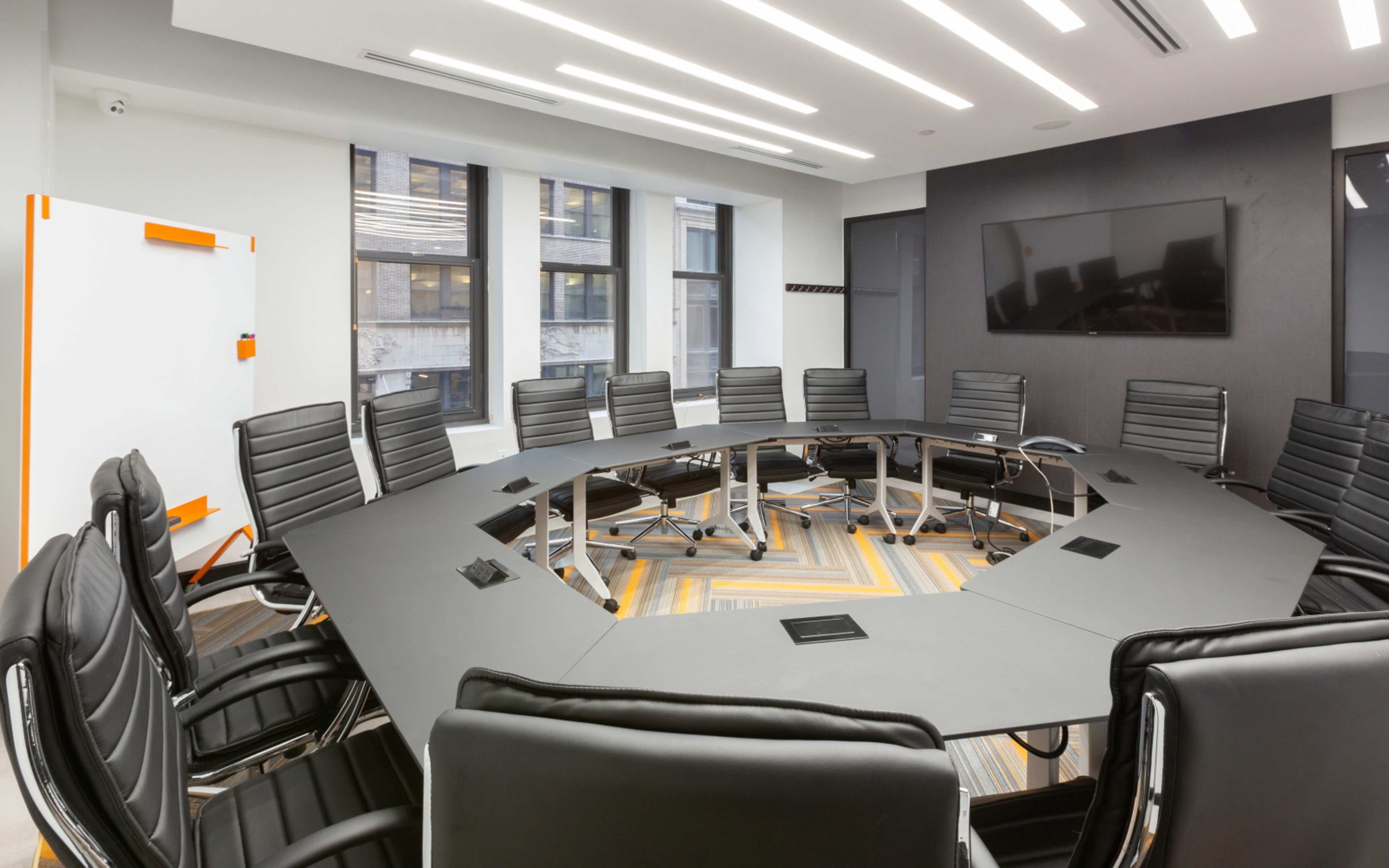 A large, round meeting table surrounded by black chairs is shown in a modern conference room with large windows and a wall-mounted screen.
