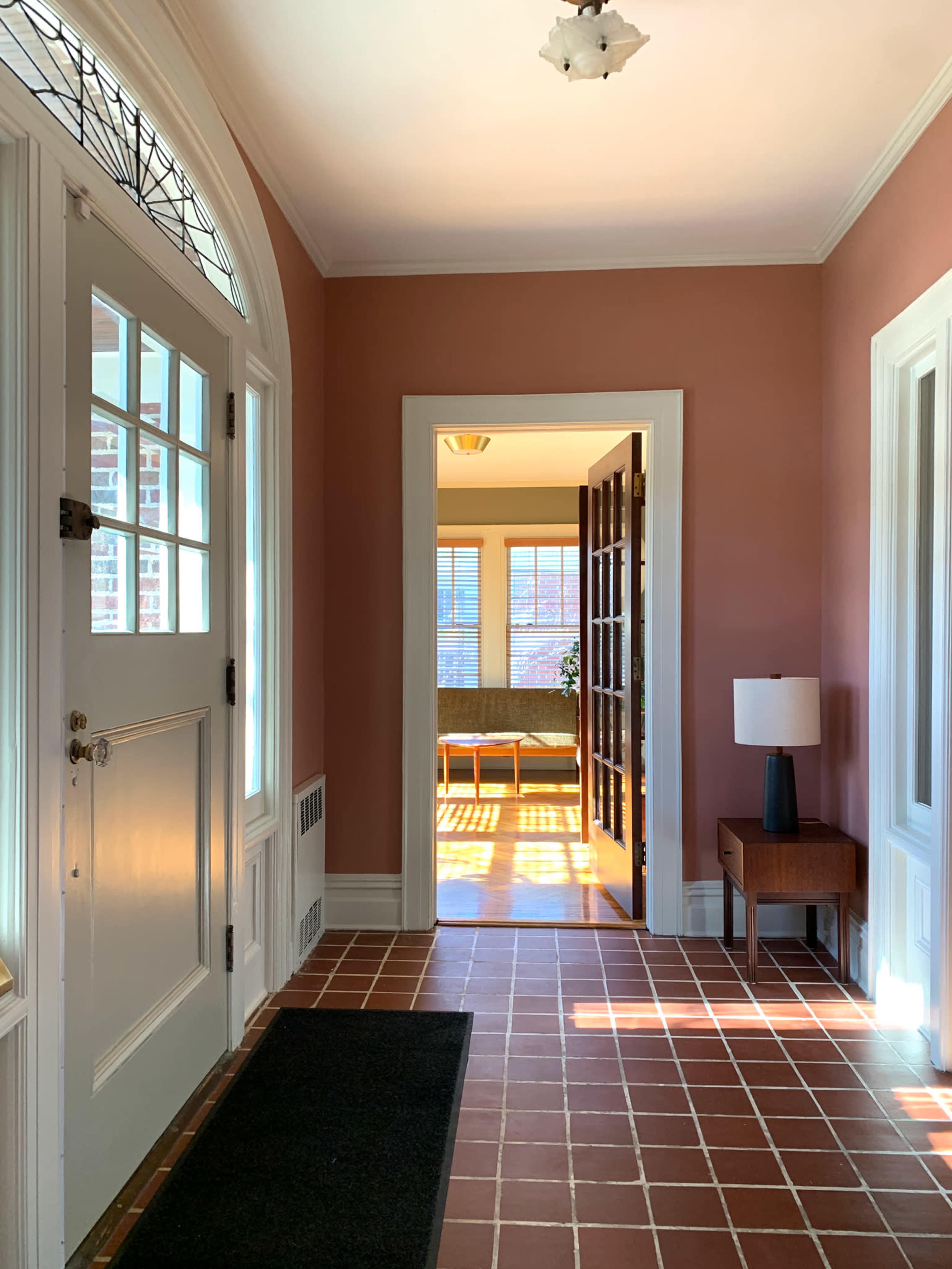 A hallway with a front door to the left, a small table with a lamp, and an open doorway leading to a sunlit room at the end.