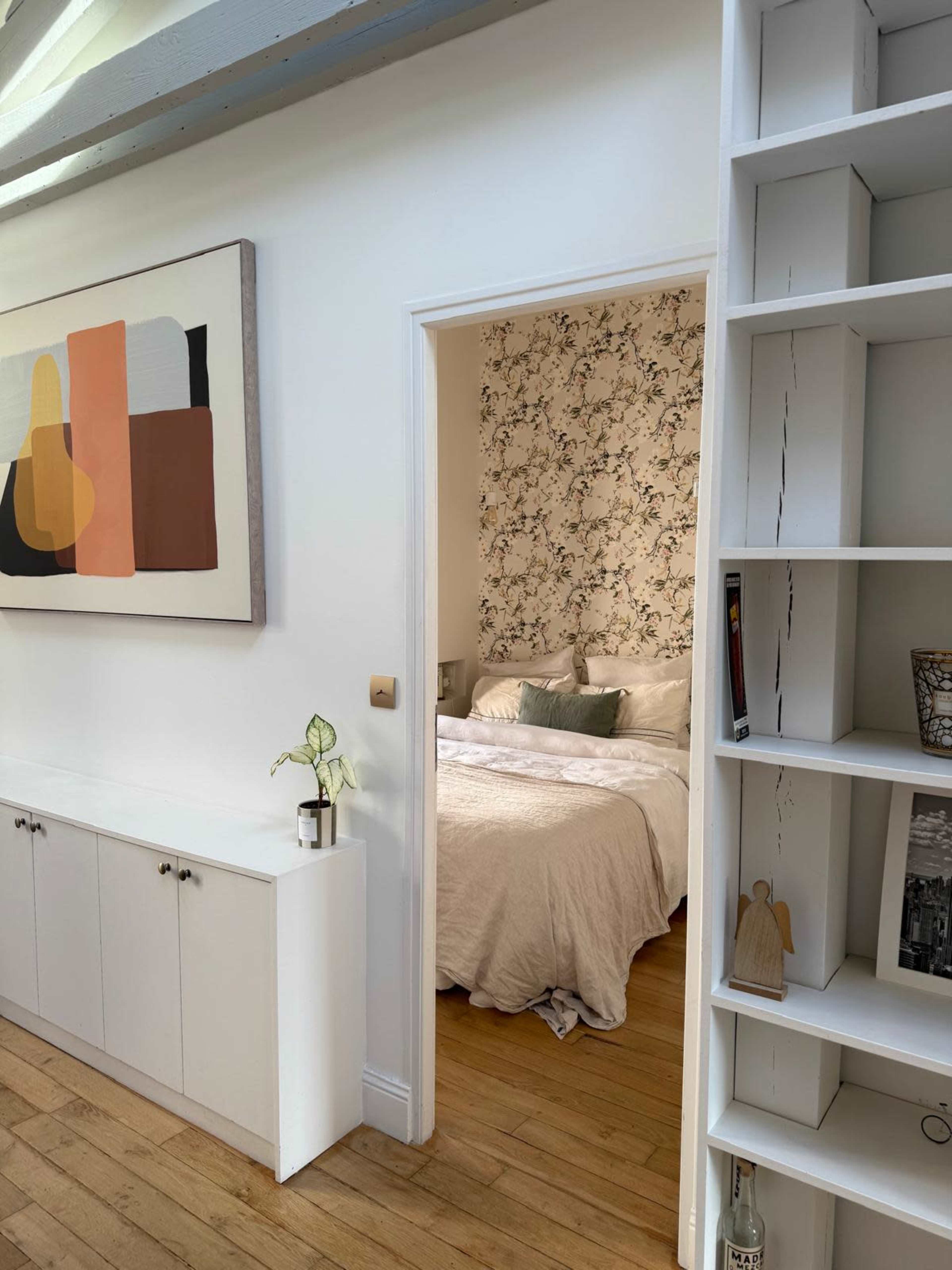 The image shows the entrance to a bedroom with floral wallpaper, visible through an open door in a brightly lit room featuring a white cabinet and decorative shelving.