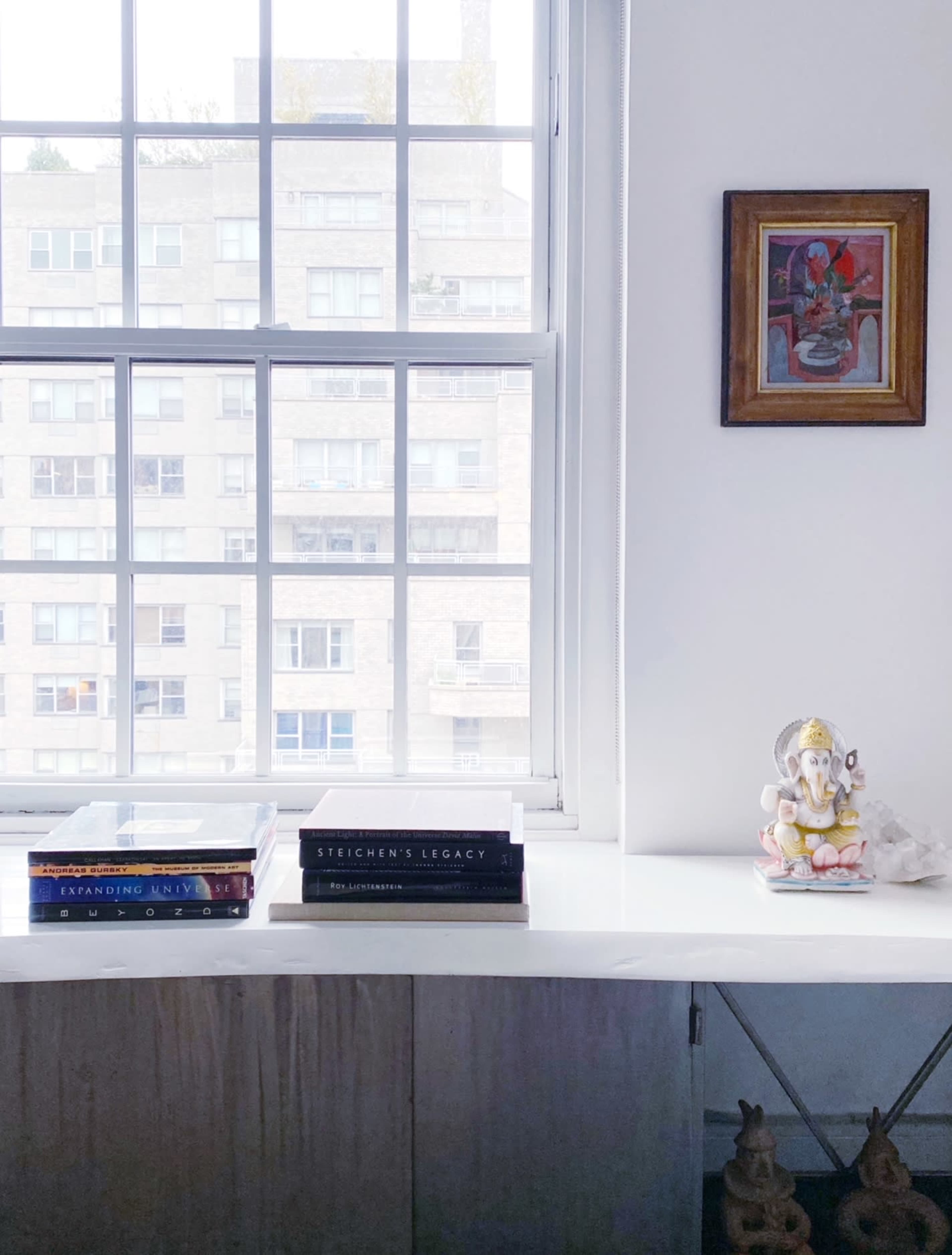 A windowsill holds stacked books and a small sculpture of Ganesha next to a framed artwork, with a view of a building visible through the window.