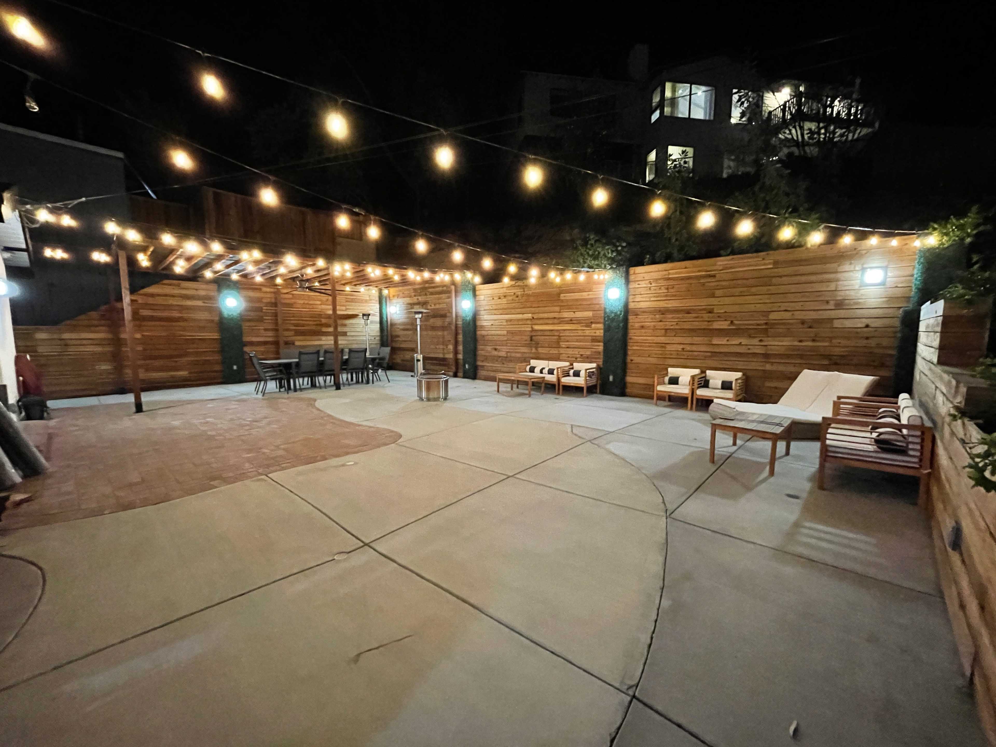 A spacious outdoor patio at night, illuminated by string lights, featuring several seating areas, a dining table, and wooden walls.