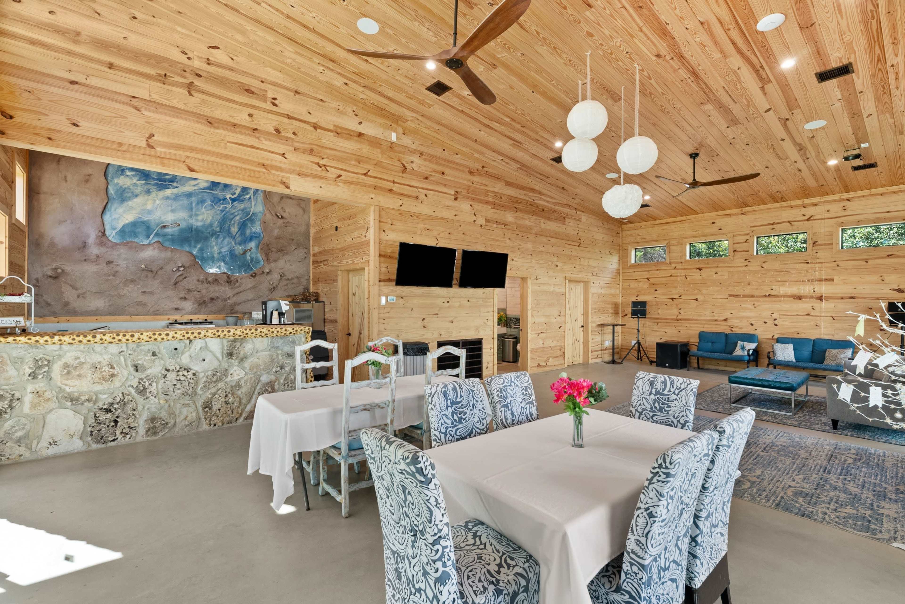 The image shows a spacious wooden interior with a stone accent wall, featuring a reception area and a dining table set with floral arrangements.