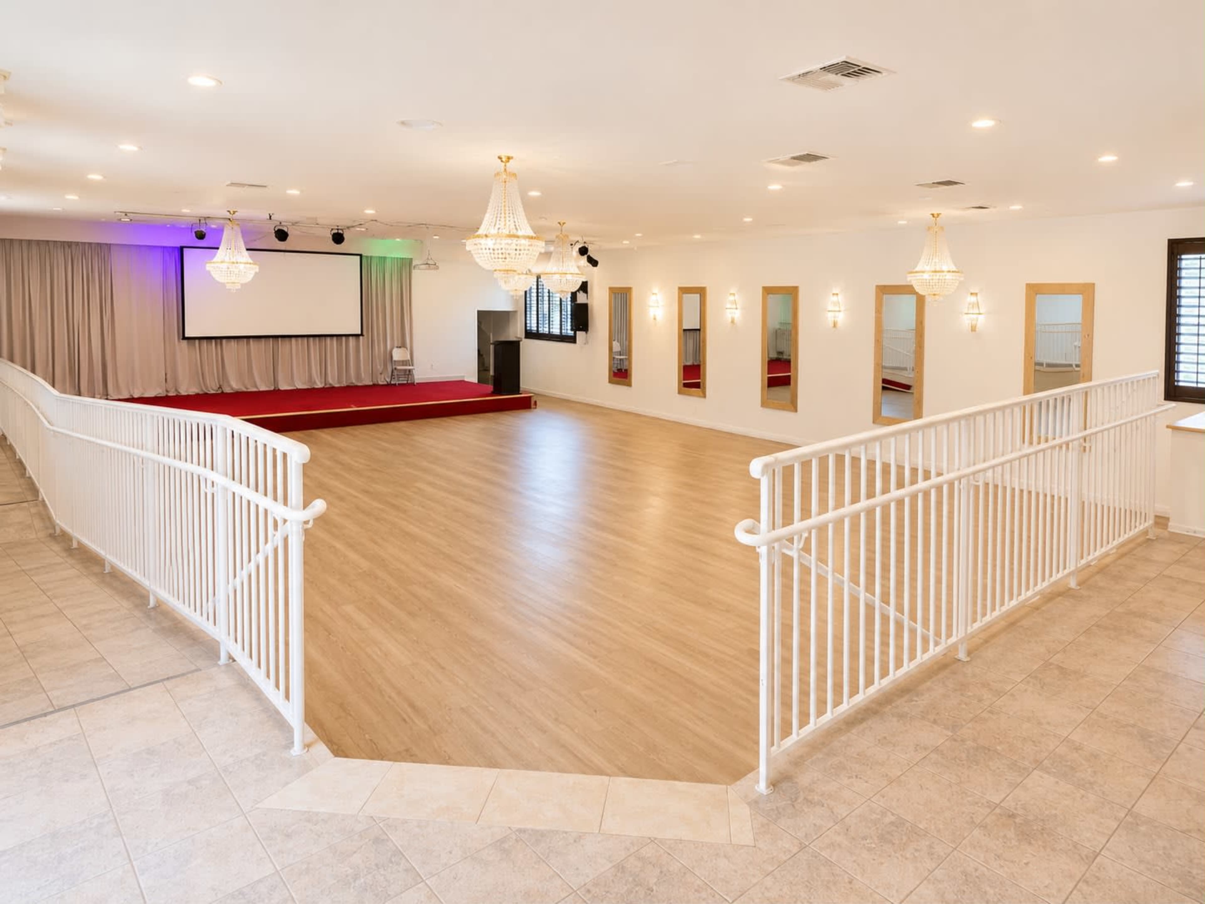 The image shows a spacious event hall with wooden flooring, chandeliers, a stage area, and mirrors lining the walls.