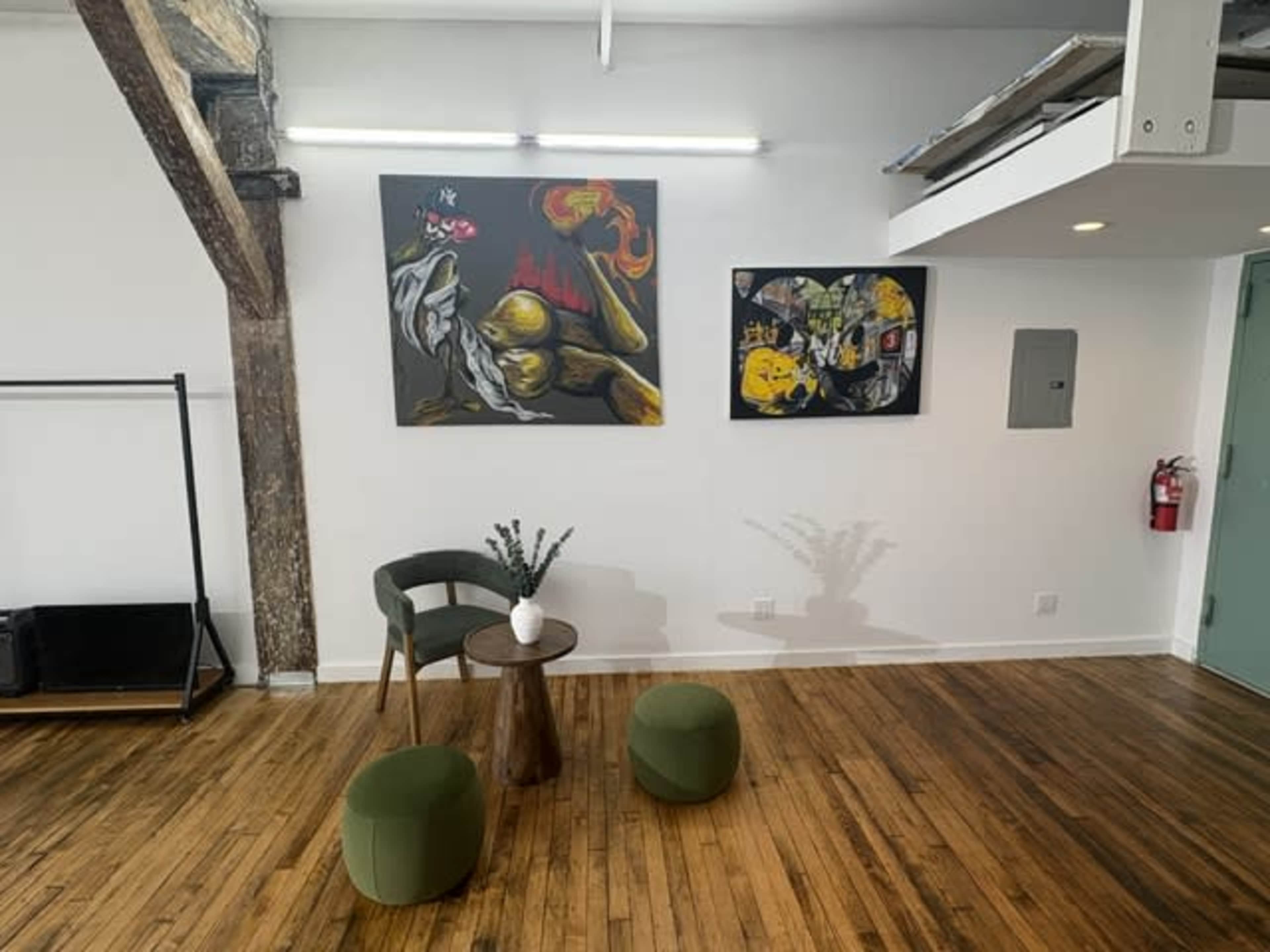 The image shows a minimalist interior with two abstract paintings on the wall, a small round table, and two upholstered stools in a room with wooden flooring and exposed beams.