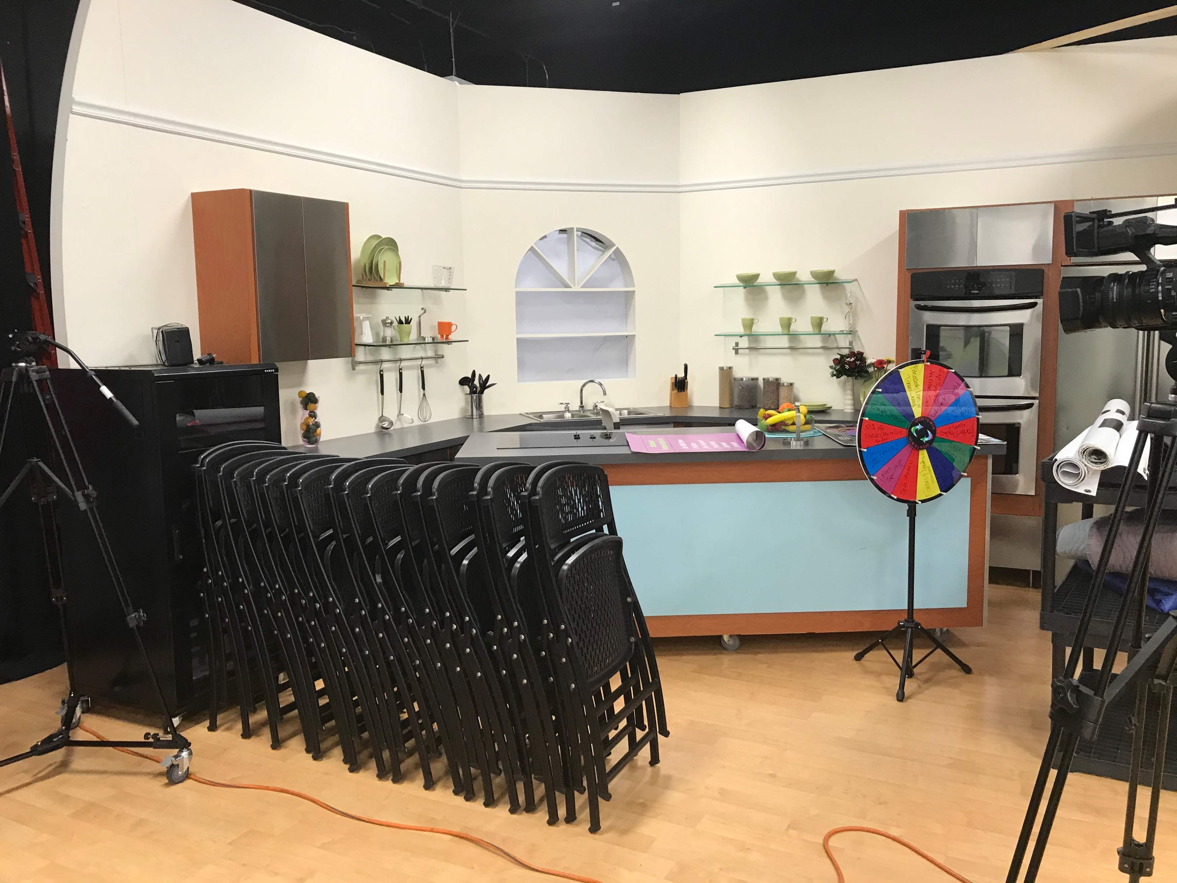 A kitchen set is prepared with a stacked arrangement of black chairs next to a countertop with various kitchen items and a colorful spinning wheel.
