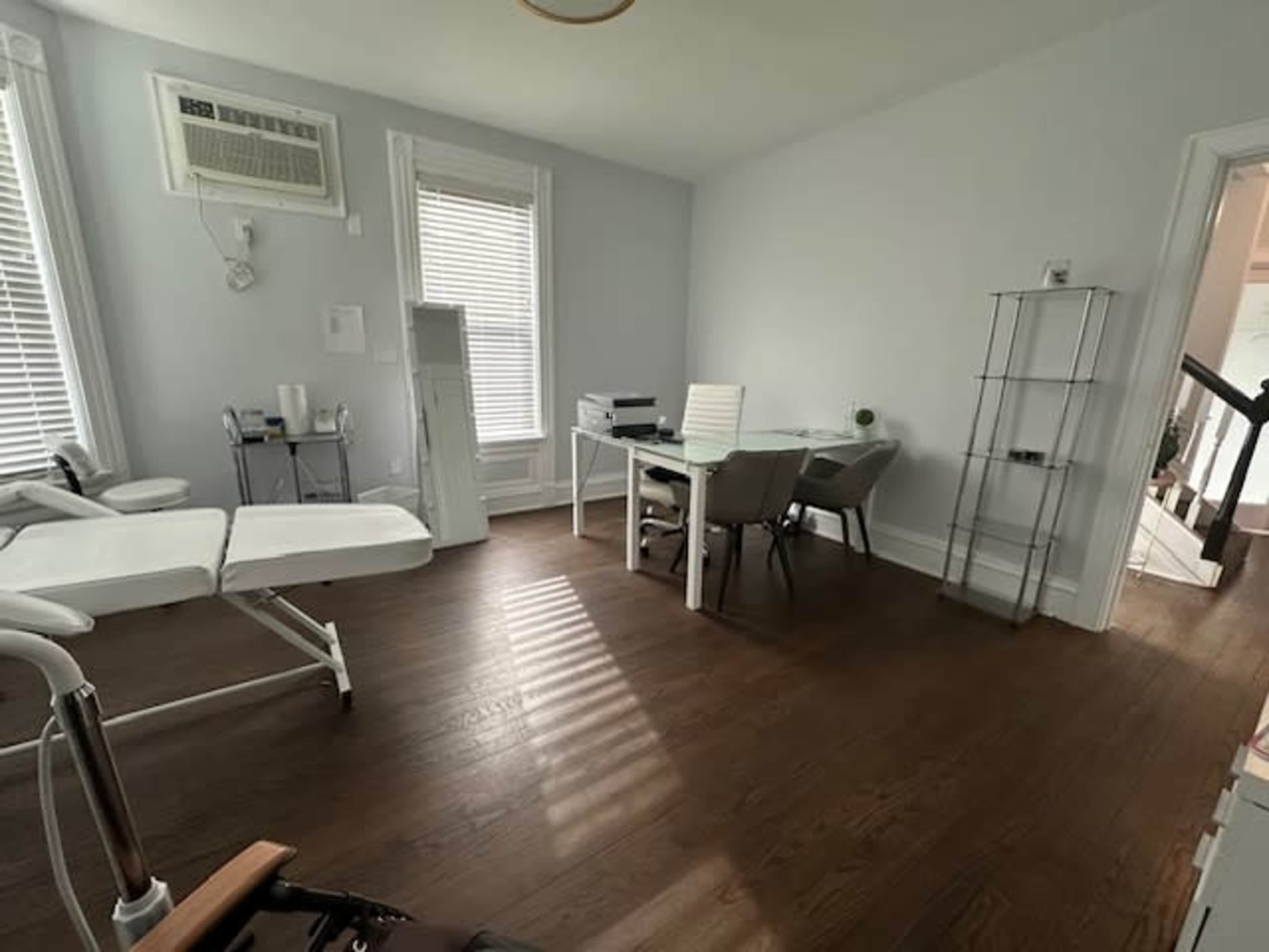 A bright, minimalist office space with a treatment table, a desk with chairs, and a shelving unit.
