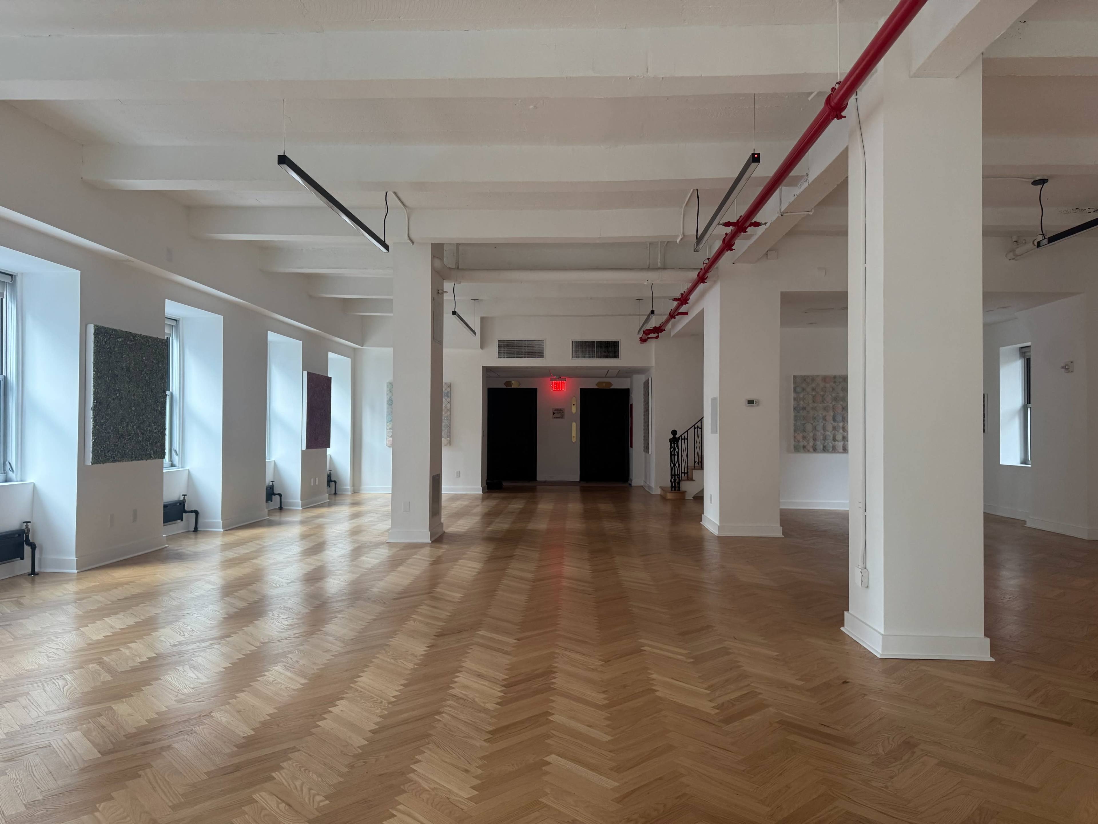The image shows a spacious, empty room with wooden herringbone flooring, large windows, and walls adorned with various artworks.