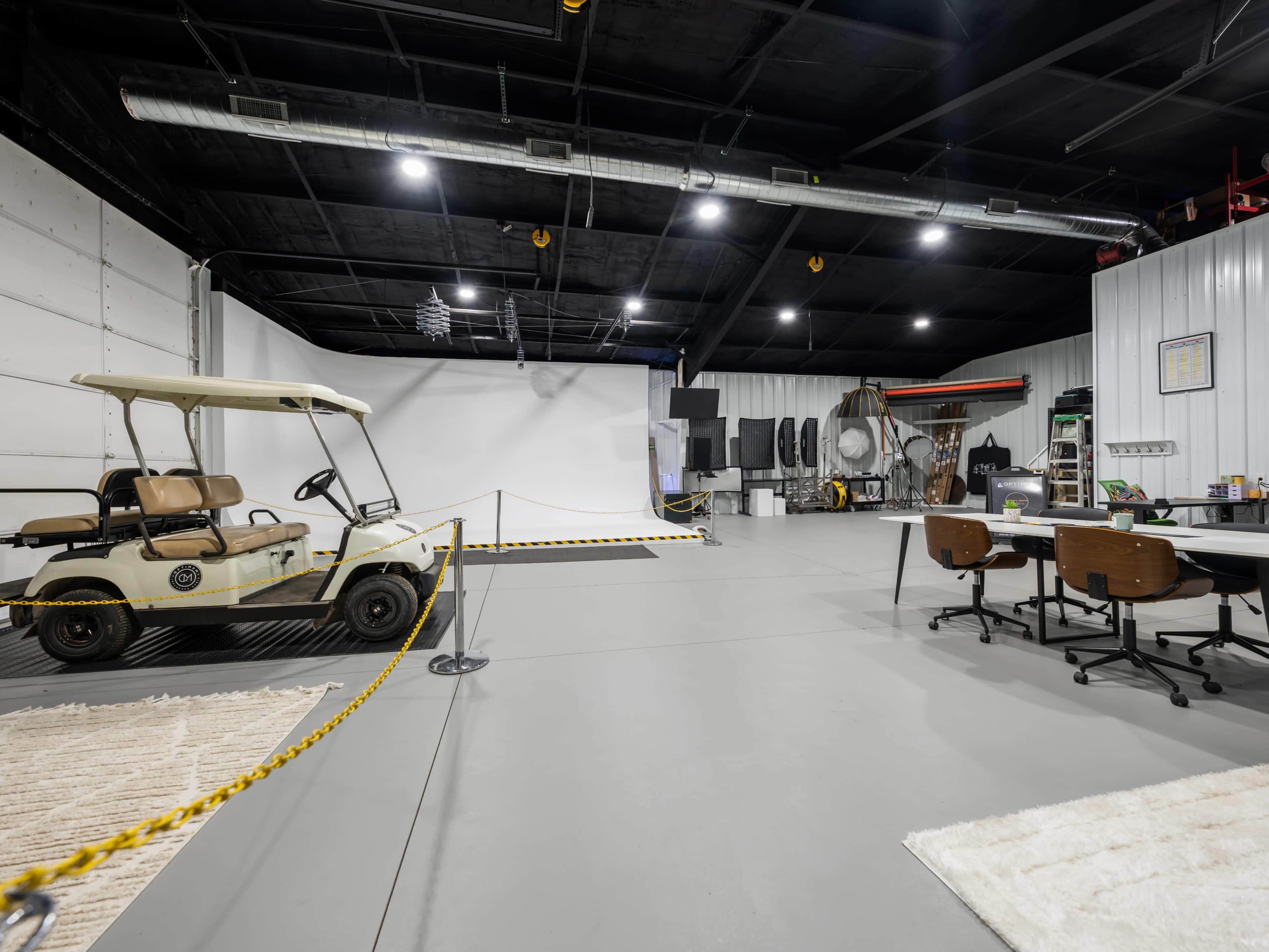 An industrial-style studio space with a golf cart parked to the left, a table with chairs on the right, and a blank backdrop at the far end.