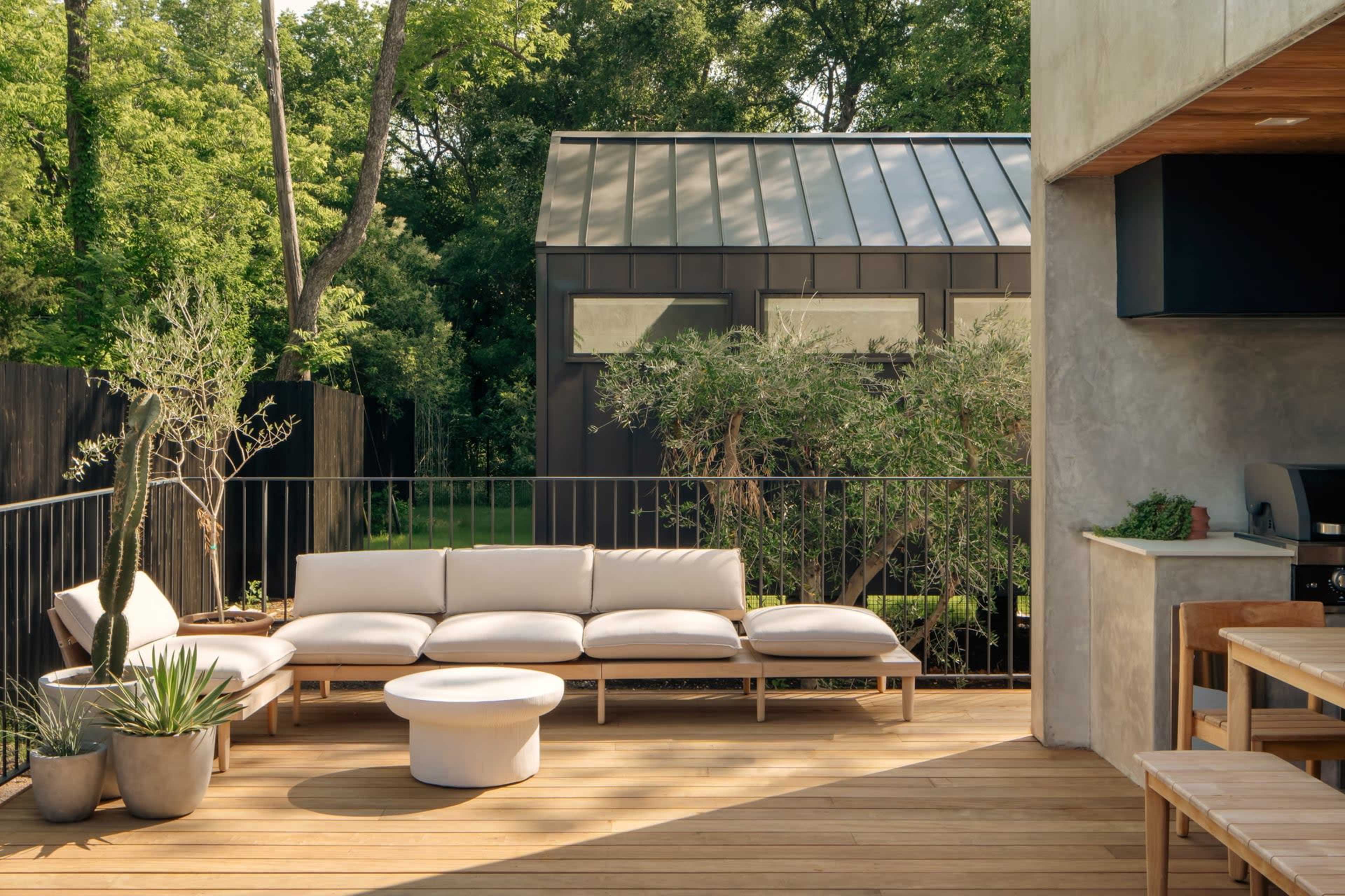 The image shows a modern outdoor patio with a light-colored sectional sofa, potted plants, and a minimalist round coffee table, set against a backdrop of greenery and a small building.