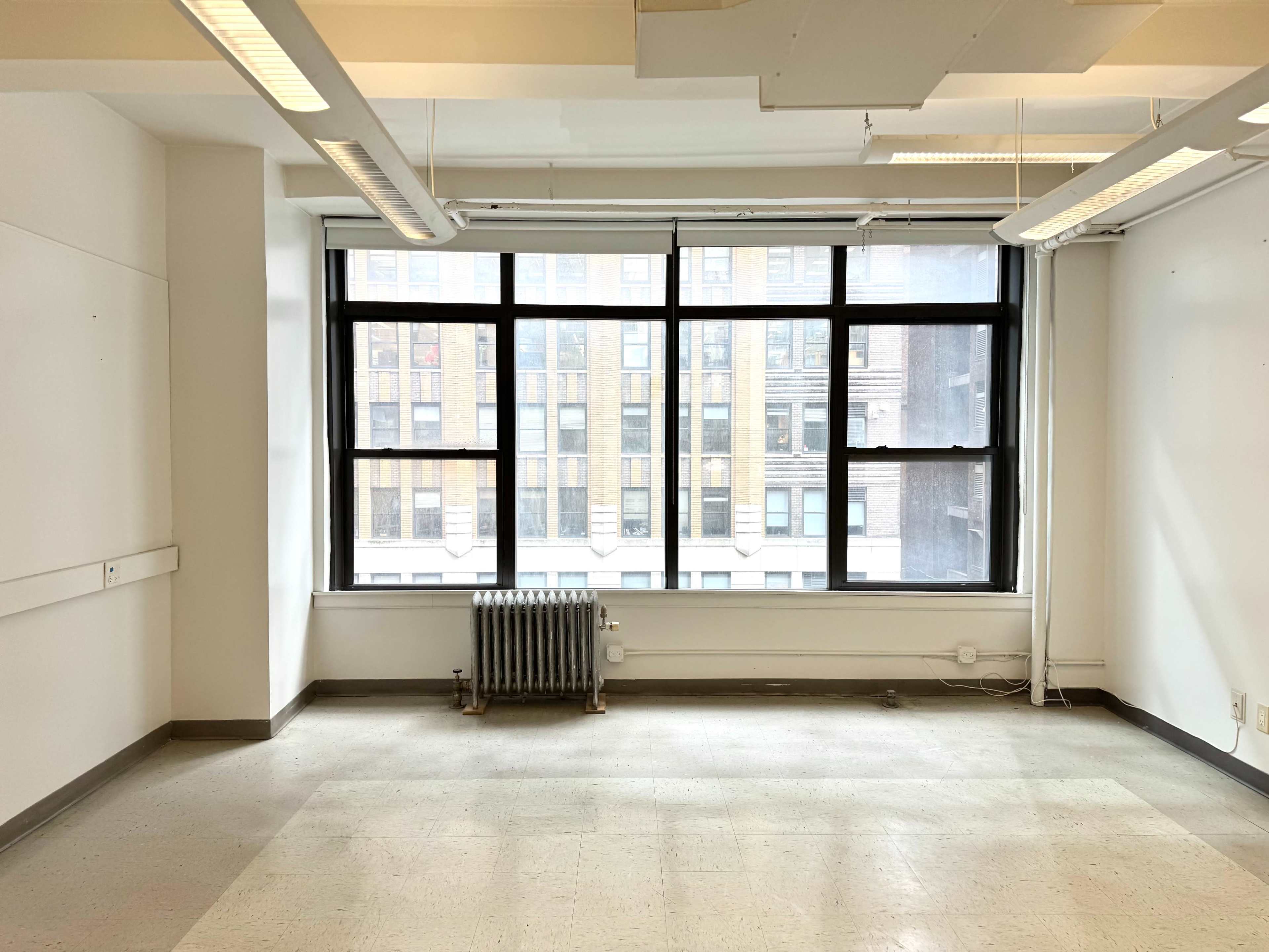 The image shows an empty room with large windows overlooking a brick building, featuring a radiator against the wall.
