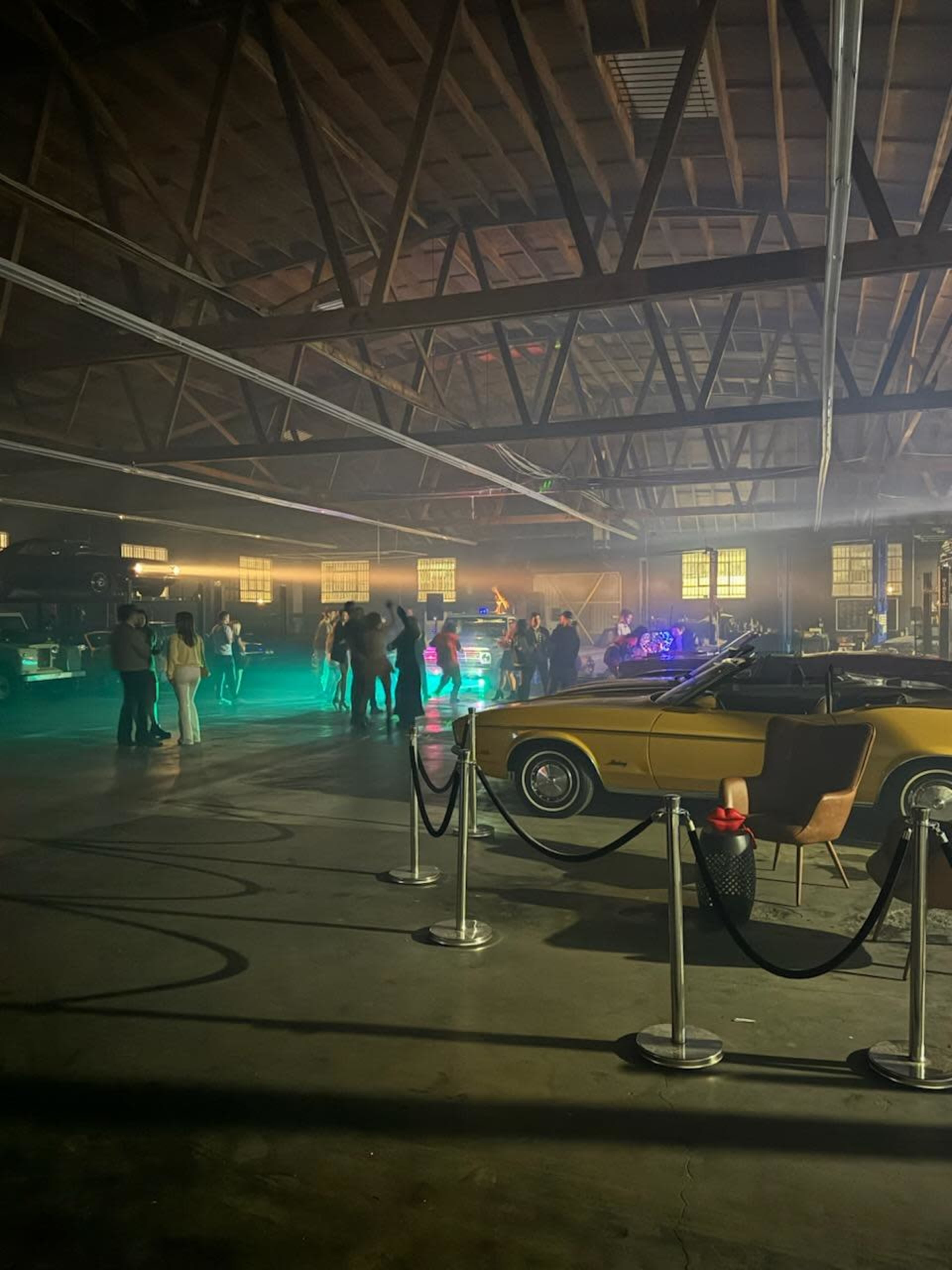 A group of people socialize in a spacious warehouse filled with classic cars and illuminated by colored lights.