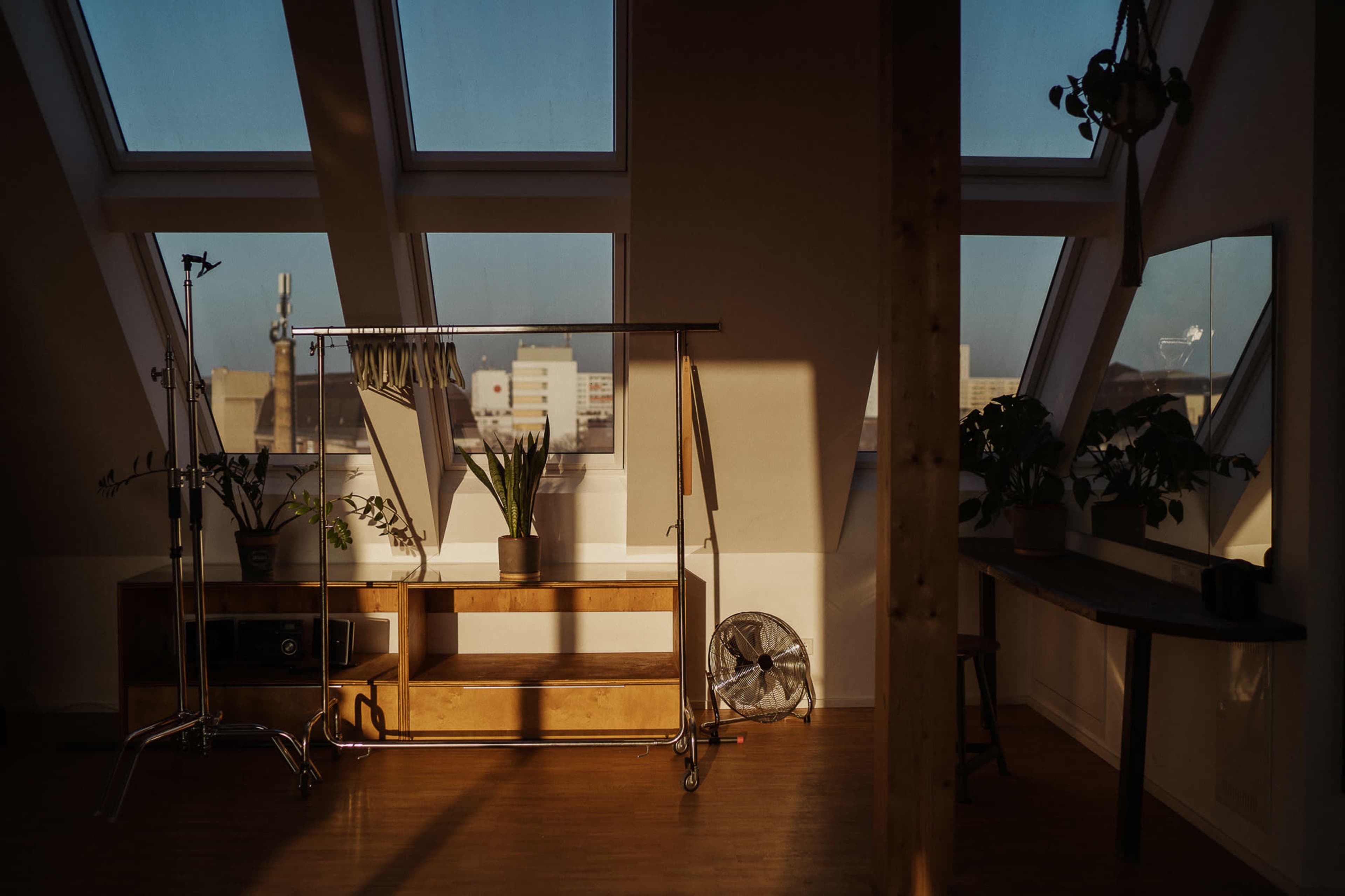 An airy attic interior with large windows, a wooden clothing rack, a shelf with plants, and a fan, casting shadows on the floor.