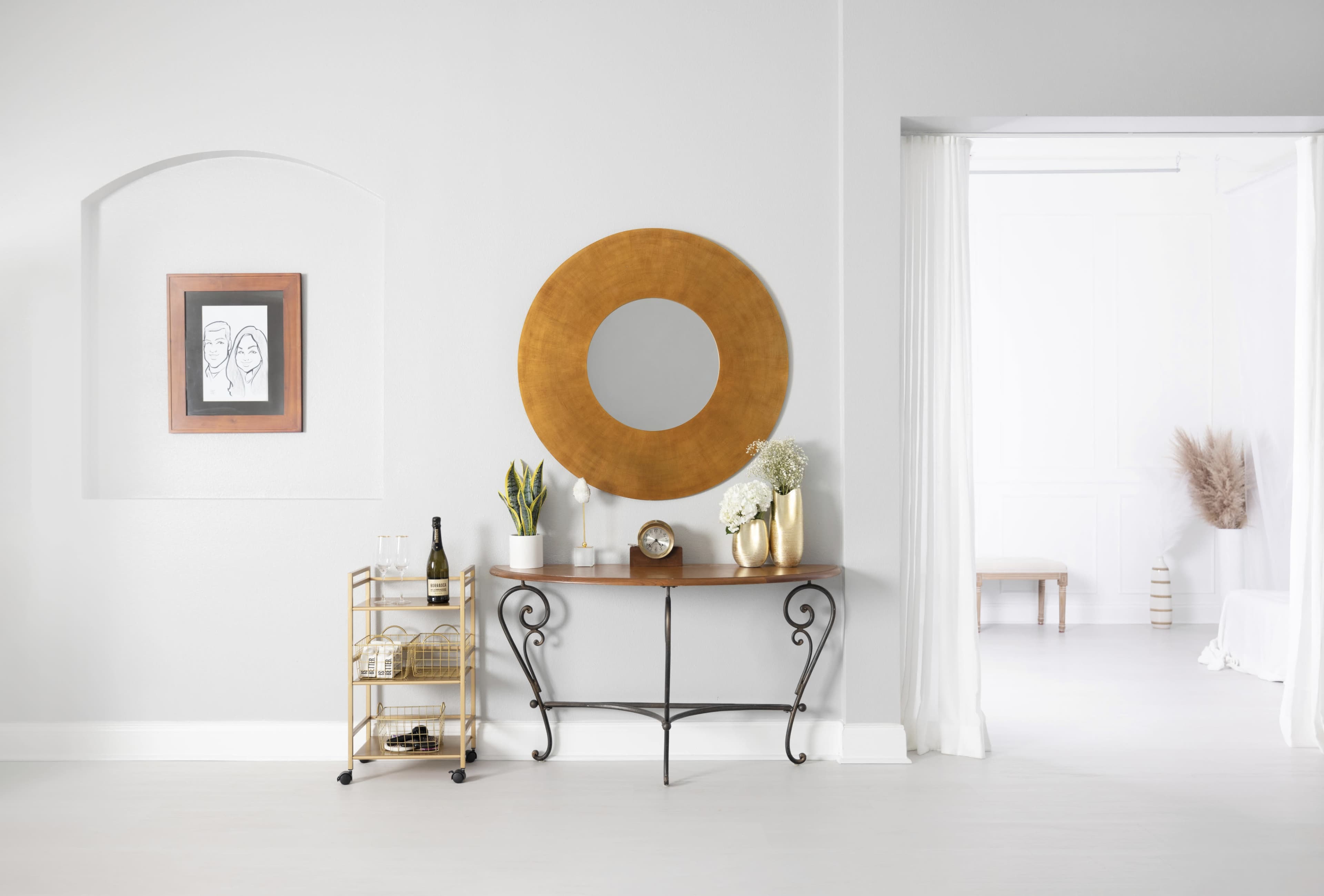 The image depicts a minimalist interior space featuring a console table against a light wall, a round mirror above it, a framed artwork, a bar cart with bottles, and a doorway leading to another room.