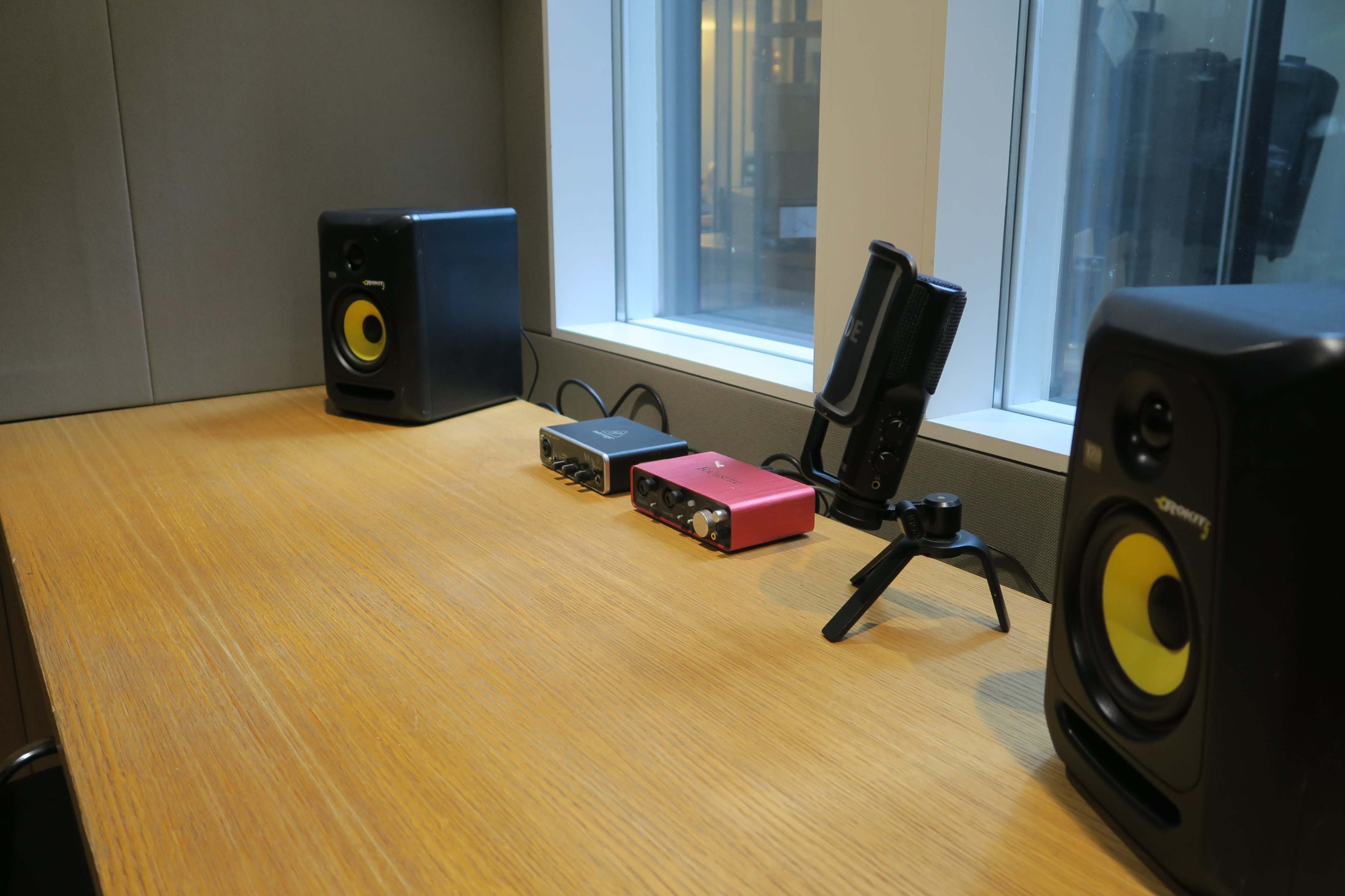 The image shows a wooden table with two audio speakers on either side, a red audio interface in the center, and a microphone mounted on a stand.