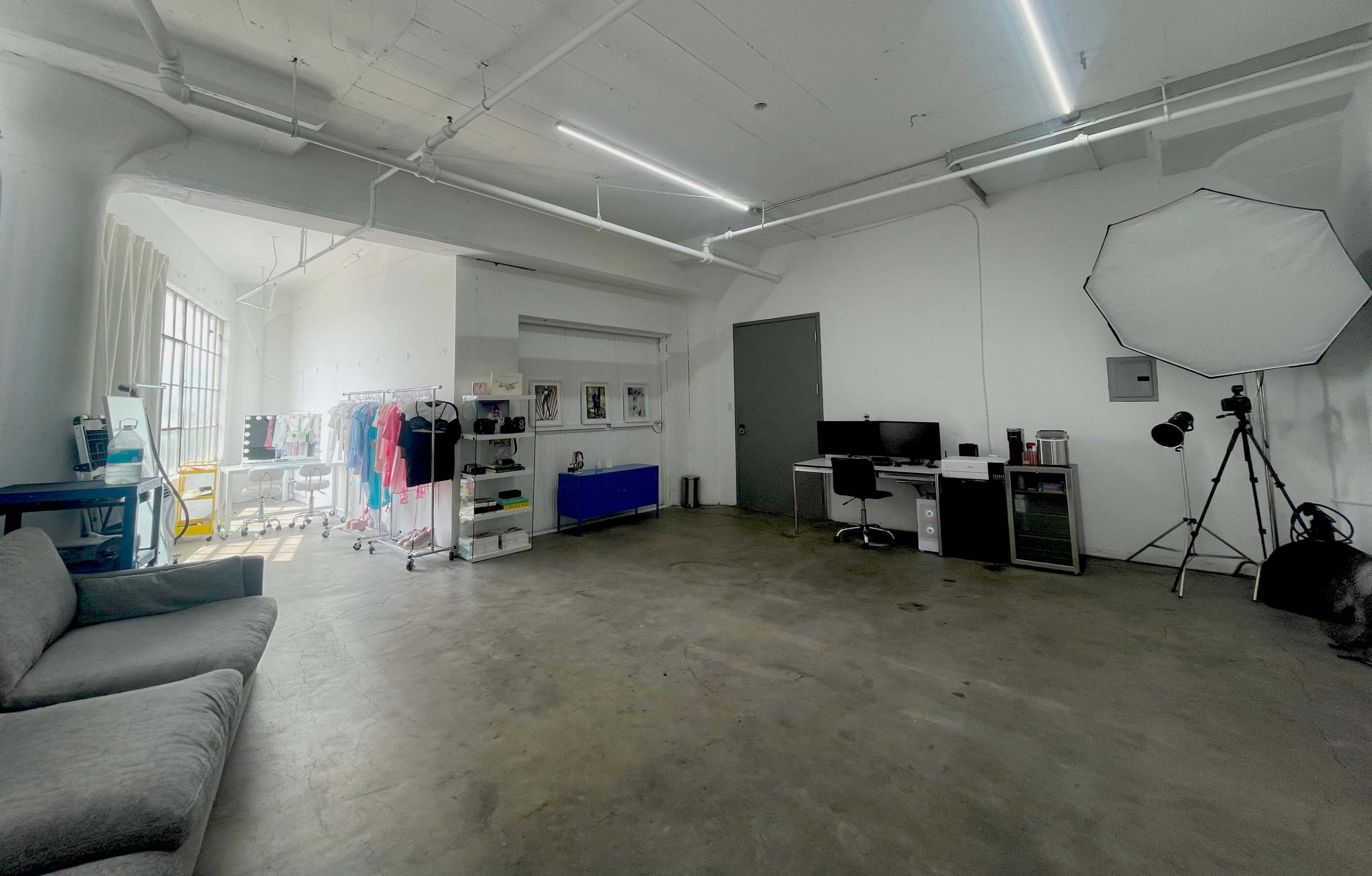 A spacious, brightly lit studio with a concrete floor, featuring a seating area, clothing racks, and a workspace equipped with a computer and photography equipment.