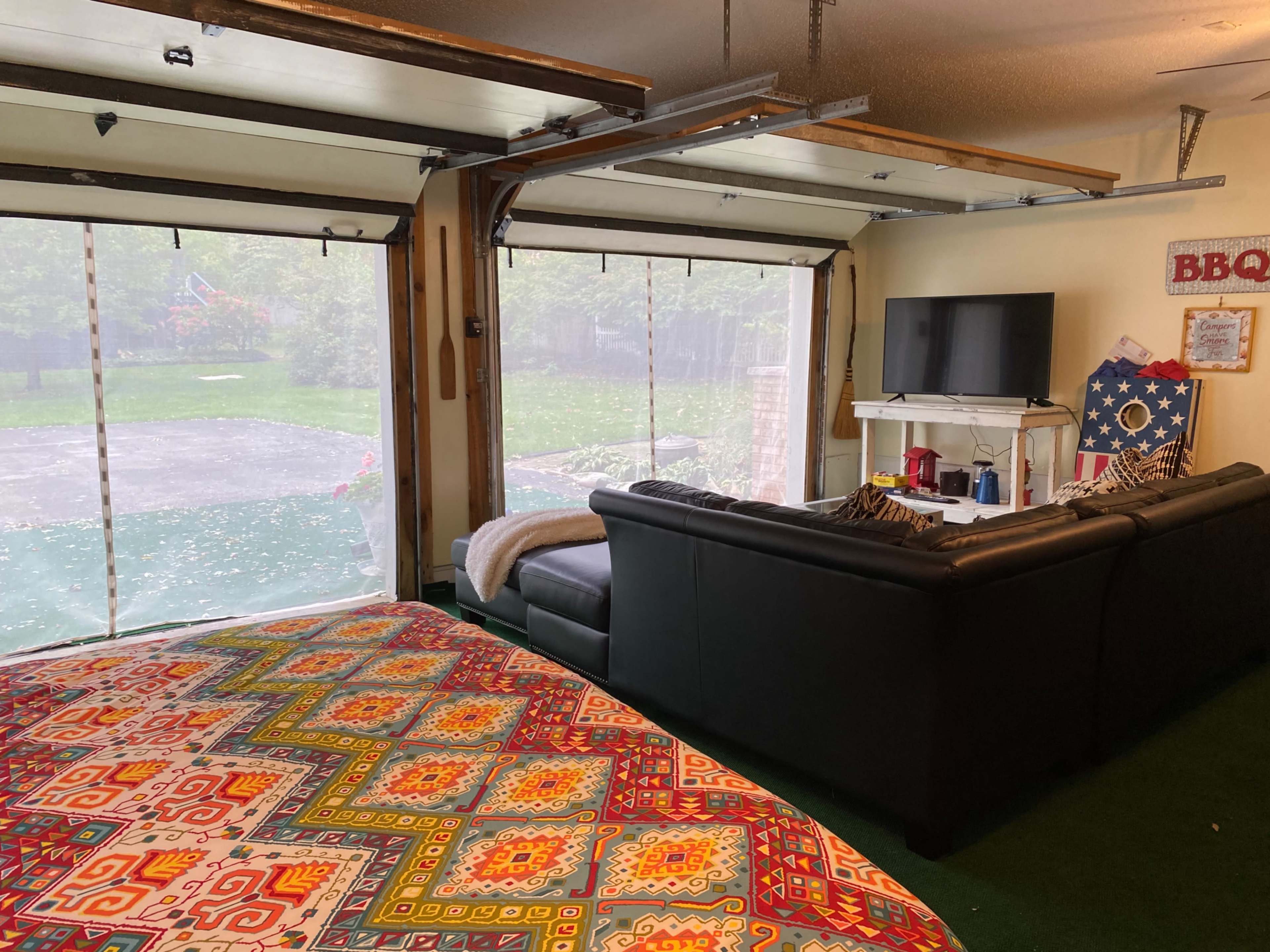 The image shows a room with a large bed covered in a colorful patterned blanket, a black leather couch, and a television, all with garage doors that provide an unobstructed view of a backyard.