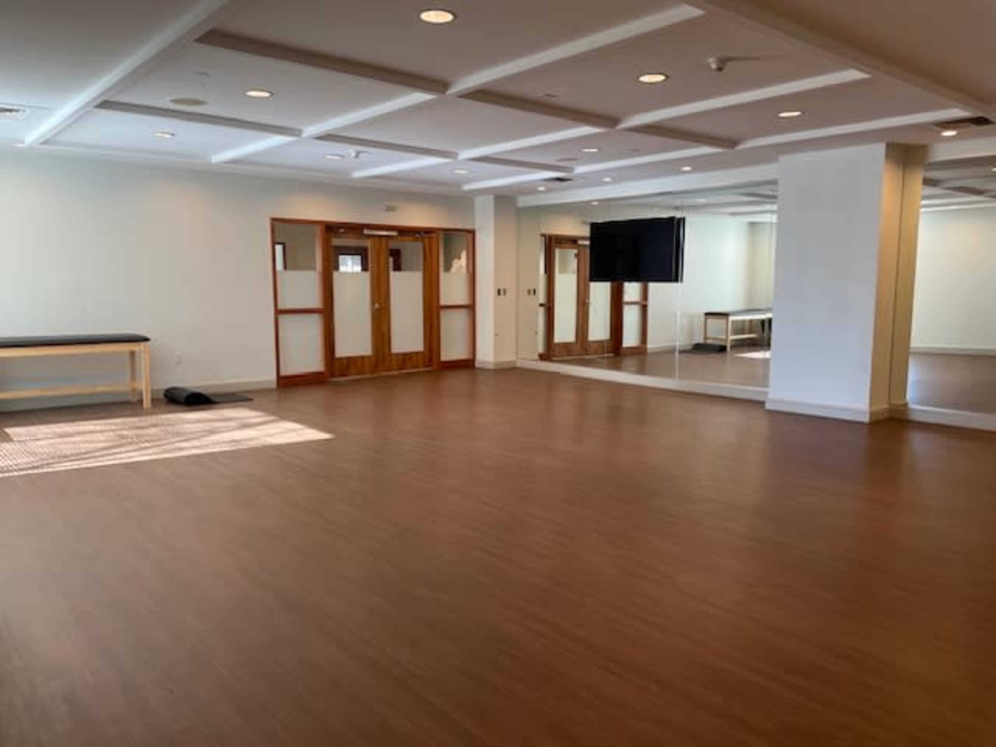 The image shows a spacious room with wooden flooring, large mirrors on one wall, and double doors leading to another area.